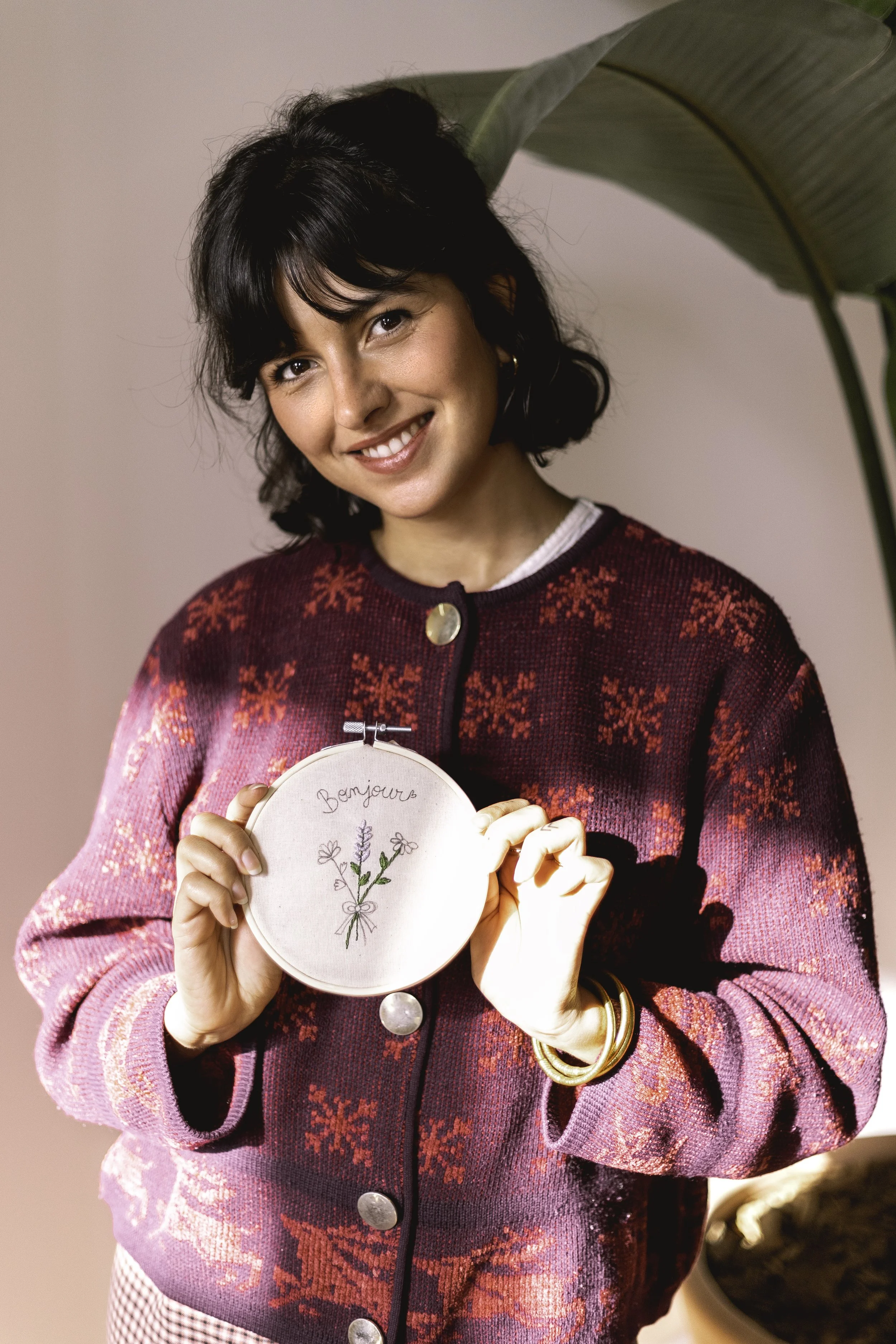 Une femme souriante tient une petite broderie avec le mot 'Bonjour' écrit dessus, entourée de fleurs."