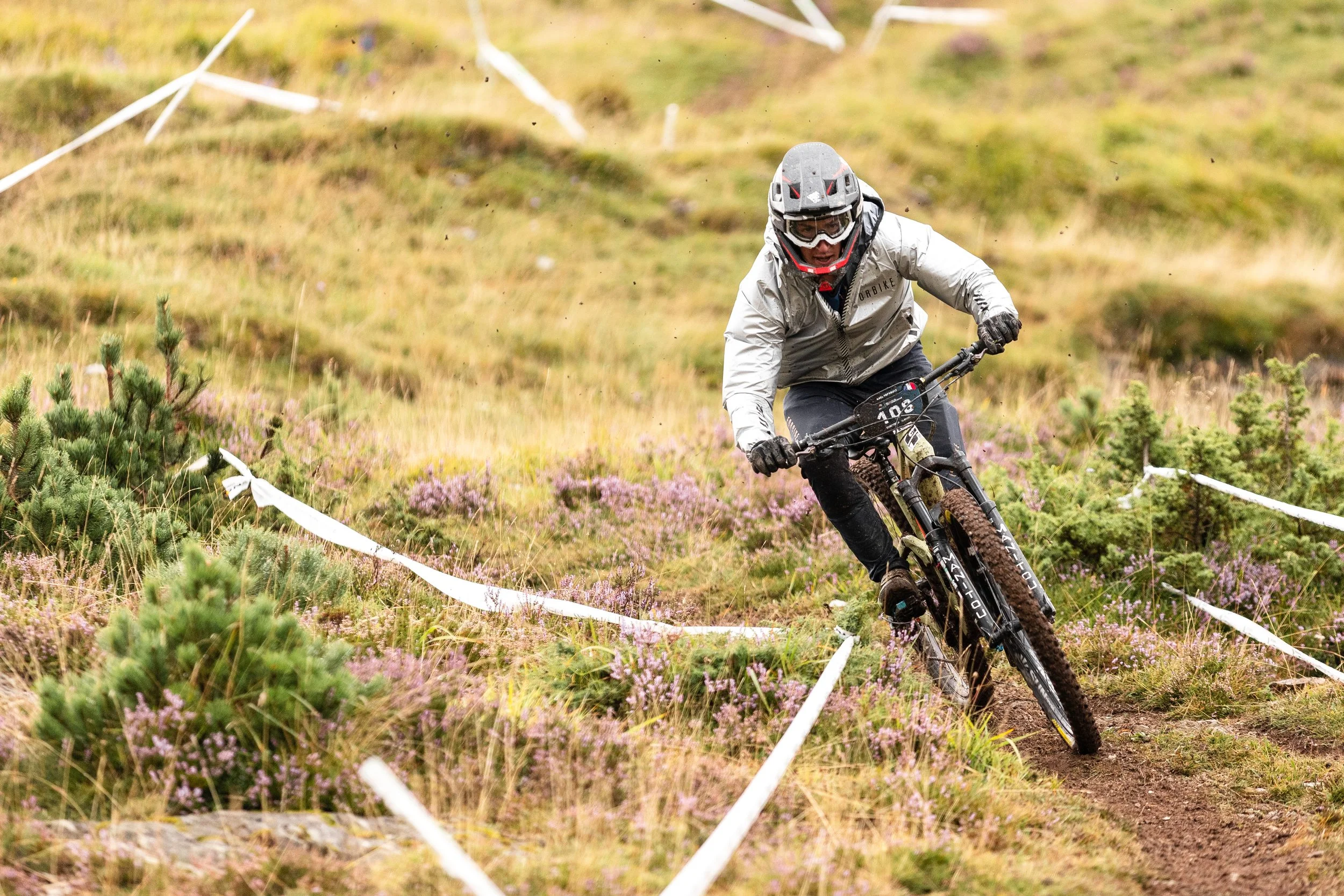 VTT de montagne en pleine course sur un sentier boueux entouré de végétation verte et de fleurs violettes, avec un casque et lunettes de protection.