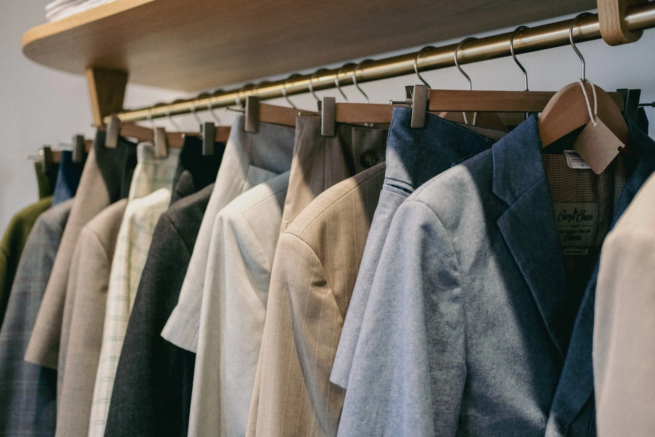 Une rangée de vestes et costumes en tissu, suspendus sur des cintres en bois, accrochés à une barre en métal, dans une garde-robe ou un magasin de vêtements.