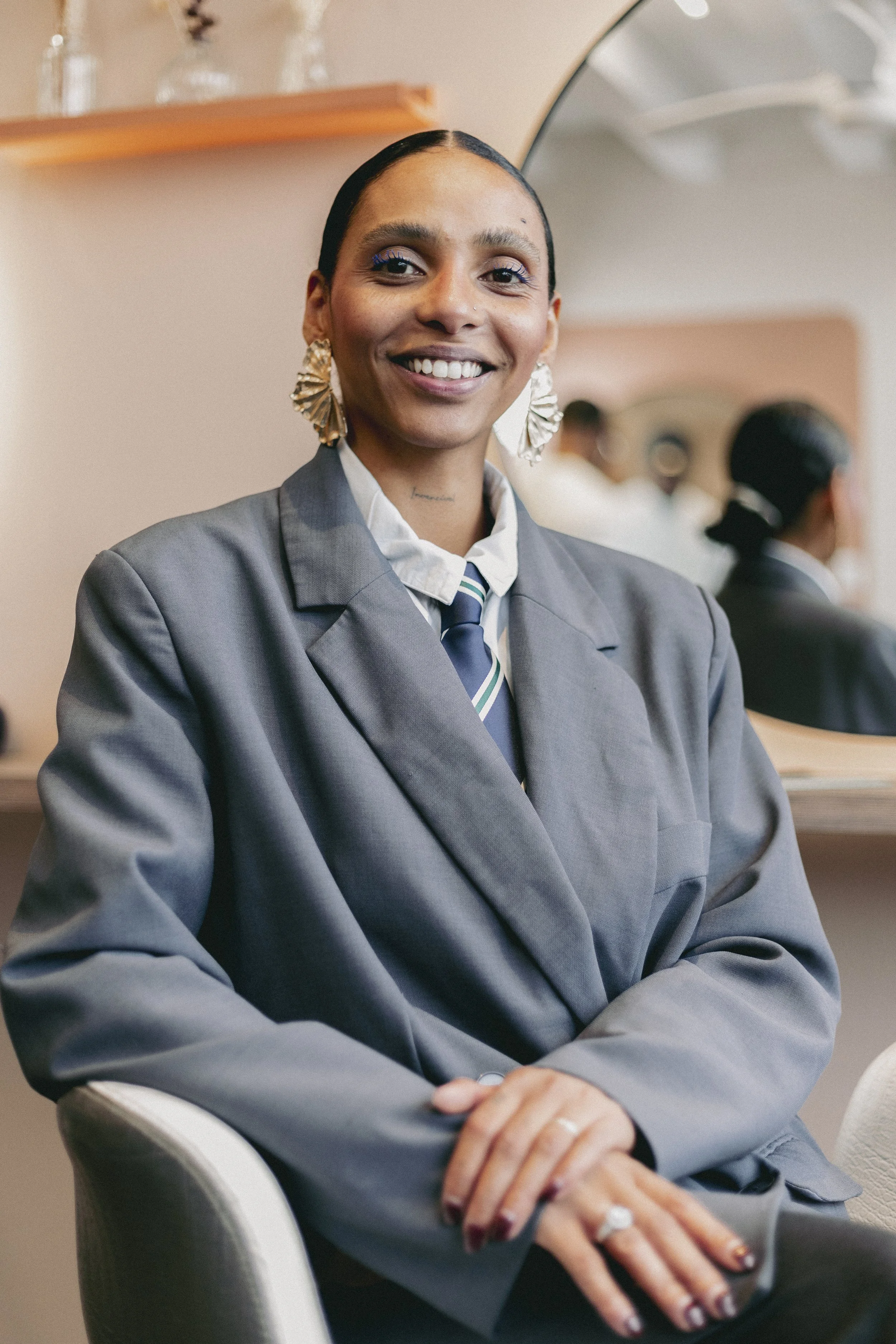 Une femme souriante portant un costume gris, une chemise blanche, un nœud papillon rayé, des boucles d'oreilles en forme de vague en or et un fond flou montrant d'autres personnes dans une salle. Elle est assise avec ses bras croisés. 