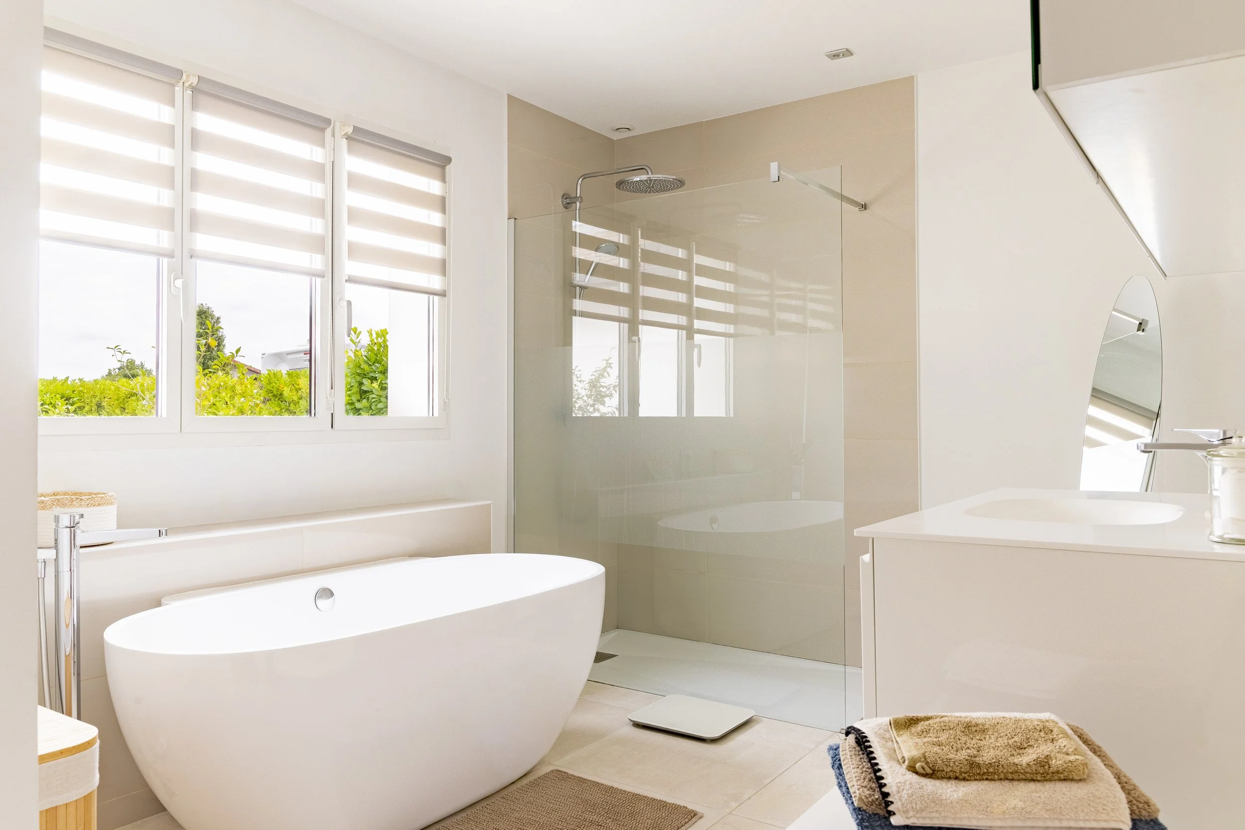 Salle de bain moderne avec une baignoire, une douche à l'italienne avec une paroi en verre, un lavabo avec un miroir, et une fenêtre avec des stores vénitiens permettant la lumière naturelle.