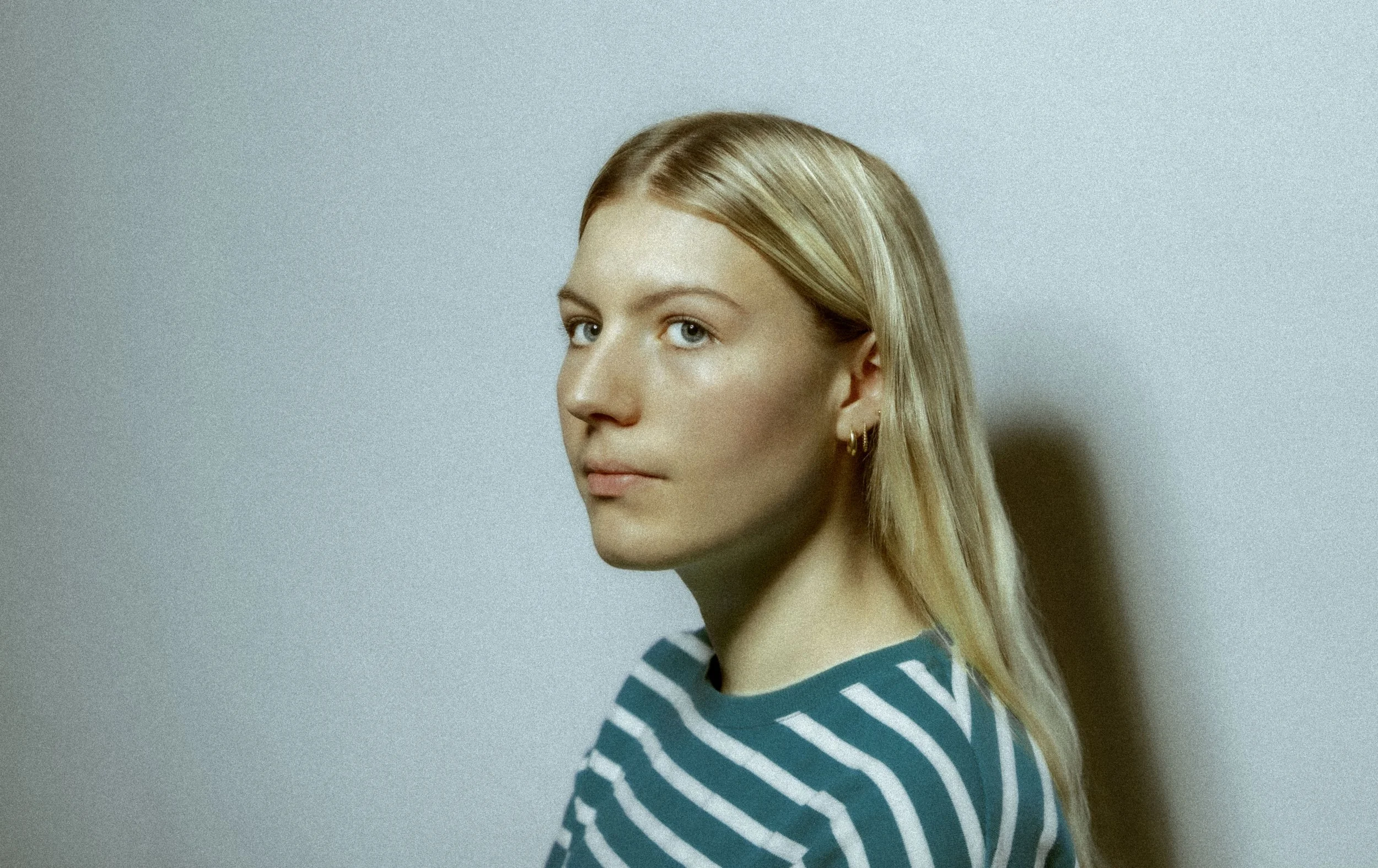 Jeune femme blonde portant un haut rayé bleu et blanc, photographiée en gros plan contre un mur clair, regardant légèrement à gauche du cadre.
