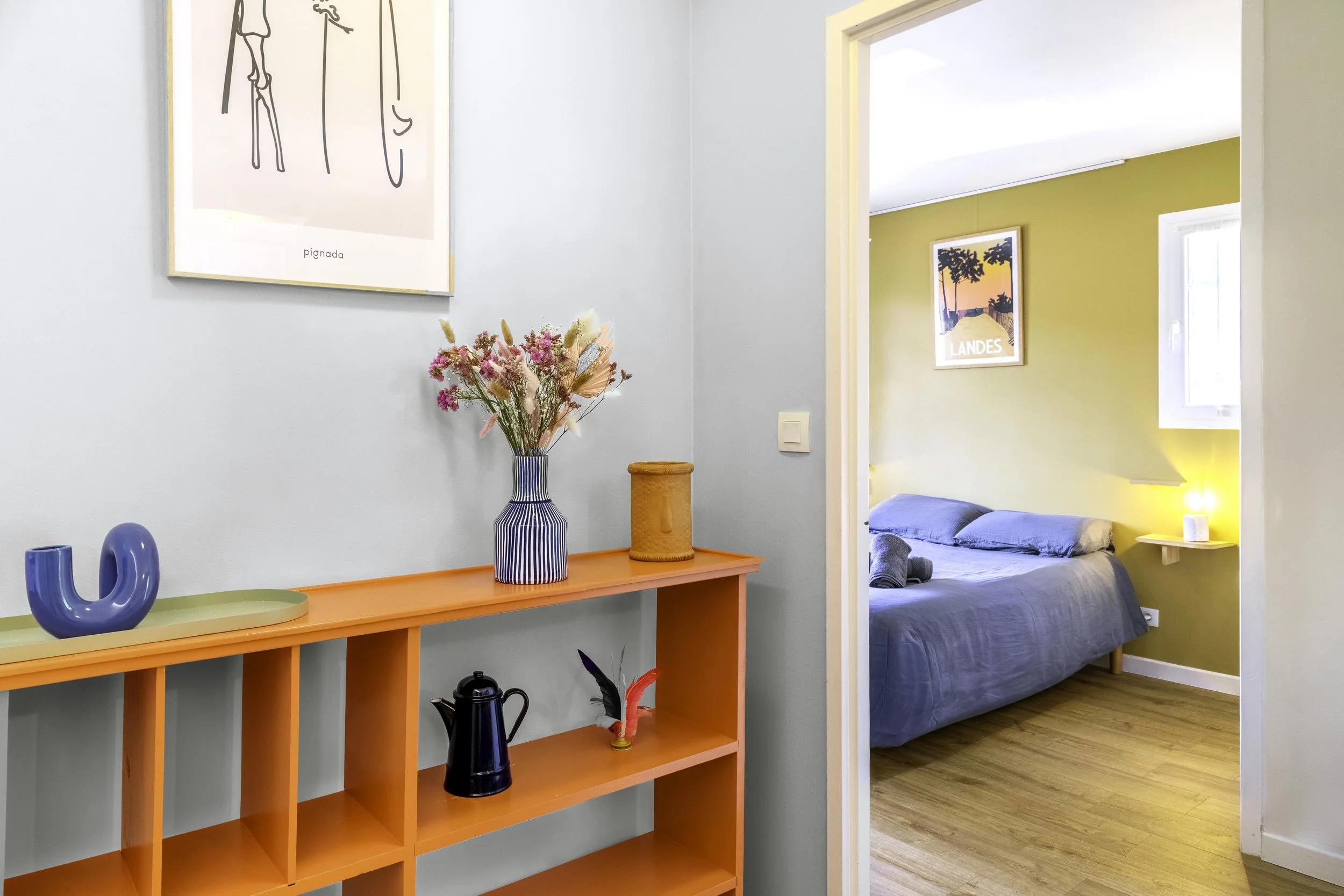 Un mur en face avec une armoire orange contenant des objets décoratifs, et une autre pièce visible avec un lit et une affiche de paysage au mur. Des fleurs dans un vase, une lampe et un objet en céramique bleue sont sur l'armoire.