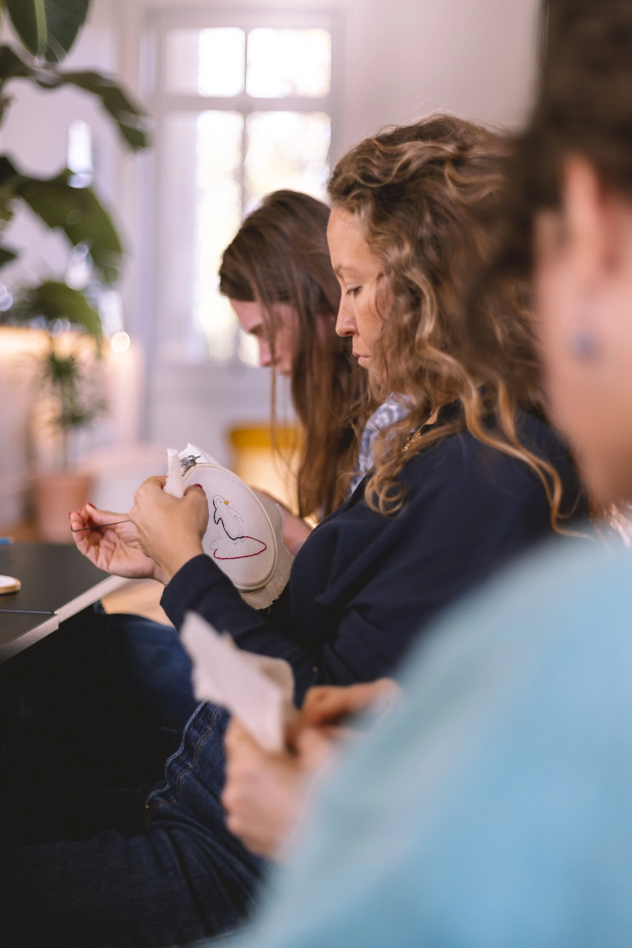 Trois femmes assises en ligne, concentrées sur leur broderie ornementale. La lumière naturelle pénètre par une fenêtre dans un intérieur lumineux.