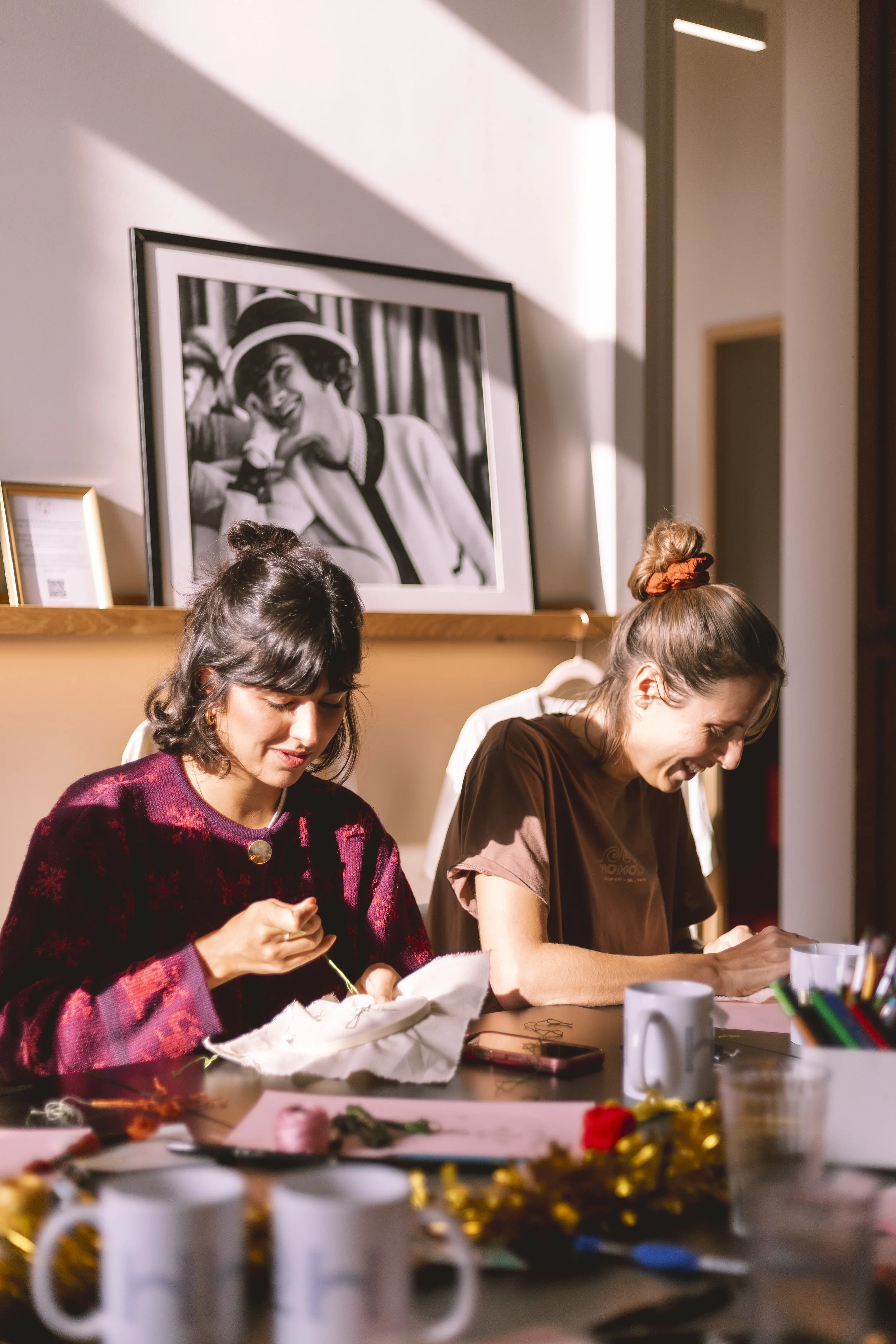 Deux femmes assises à une table créant un projet d'art ou de bricolage, avec diverses fournitures sur la table, dans un environnement intérieur bien éclairé, avec une œuvre d'art en noir et blanc en arrière-plan.