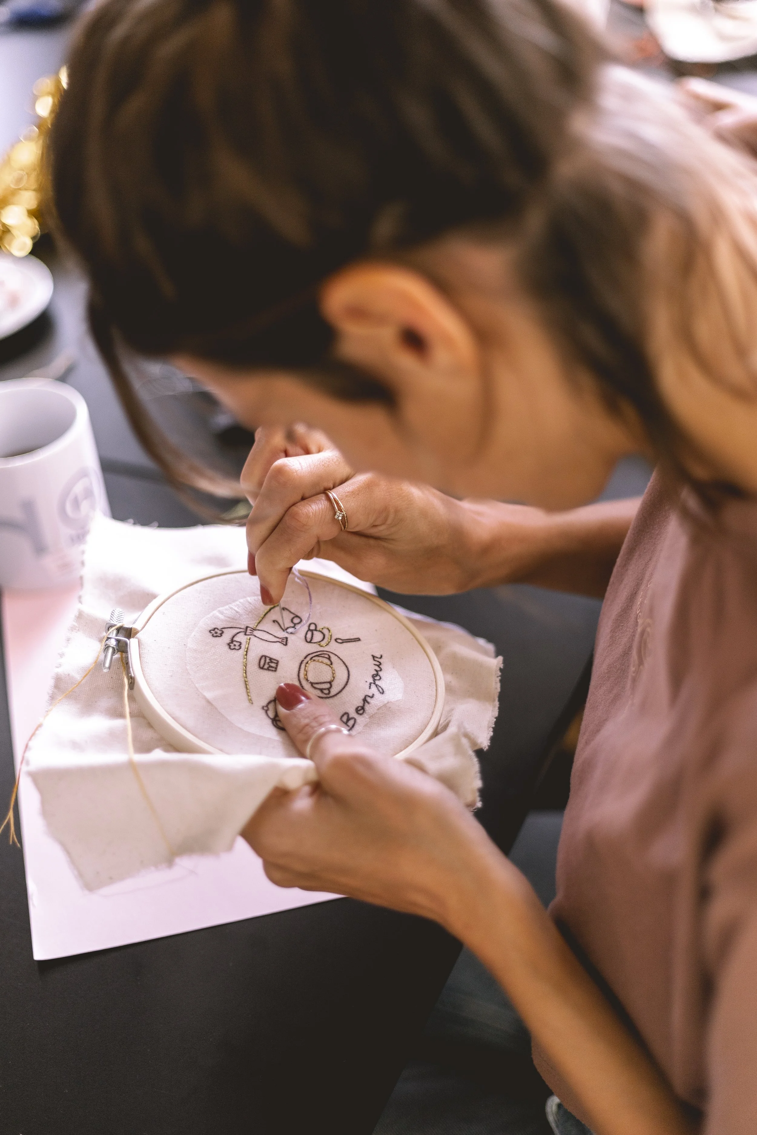 Une personne faisant de la broderie sur un tissu blanc, avec un motif coloré et le mot "Bonheur" écrit. La personne porte une bague et travaille sur une table avec des tasses et des assiettes à proximité.
