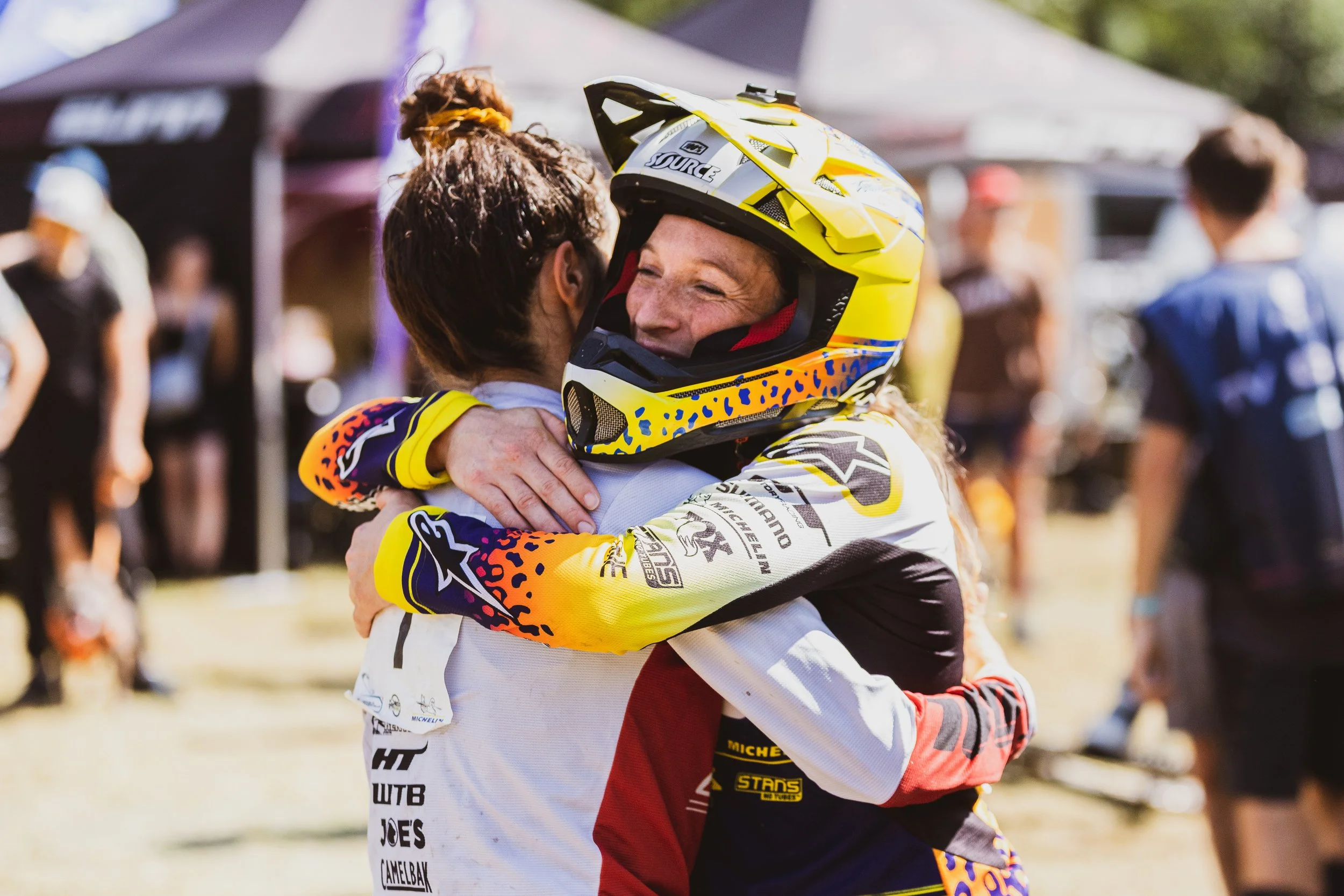Deux personnes en tenue de motocross, une portant un casque jaune, s'embrassent ou se félicitent lors d'une compétition ou d'un événement sportif en plein air.