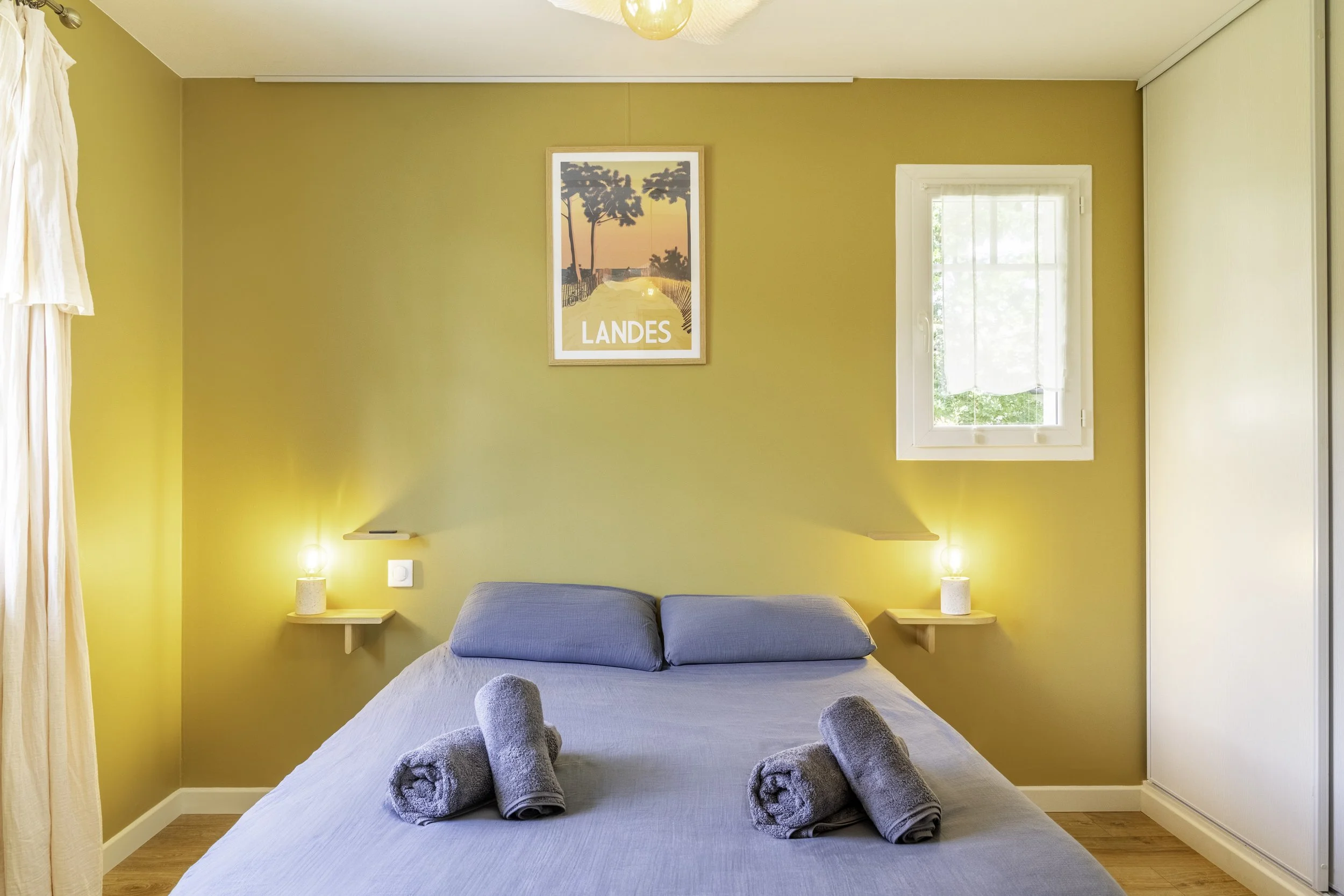 Chambre avec lit, deux serviettes roulées en forme d'animaux sur le lit, une fenêtre, tableau avec palmier et le mot 'Landes' sur le mur