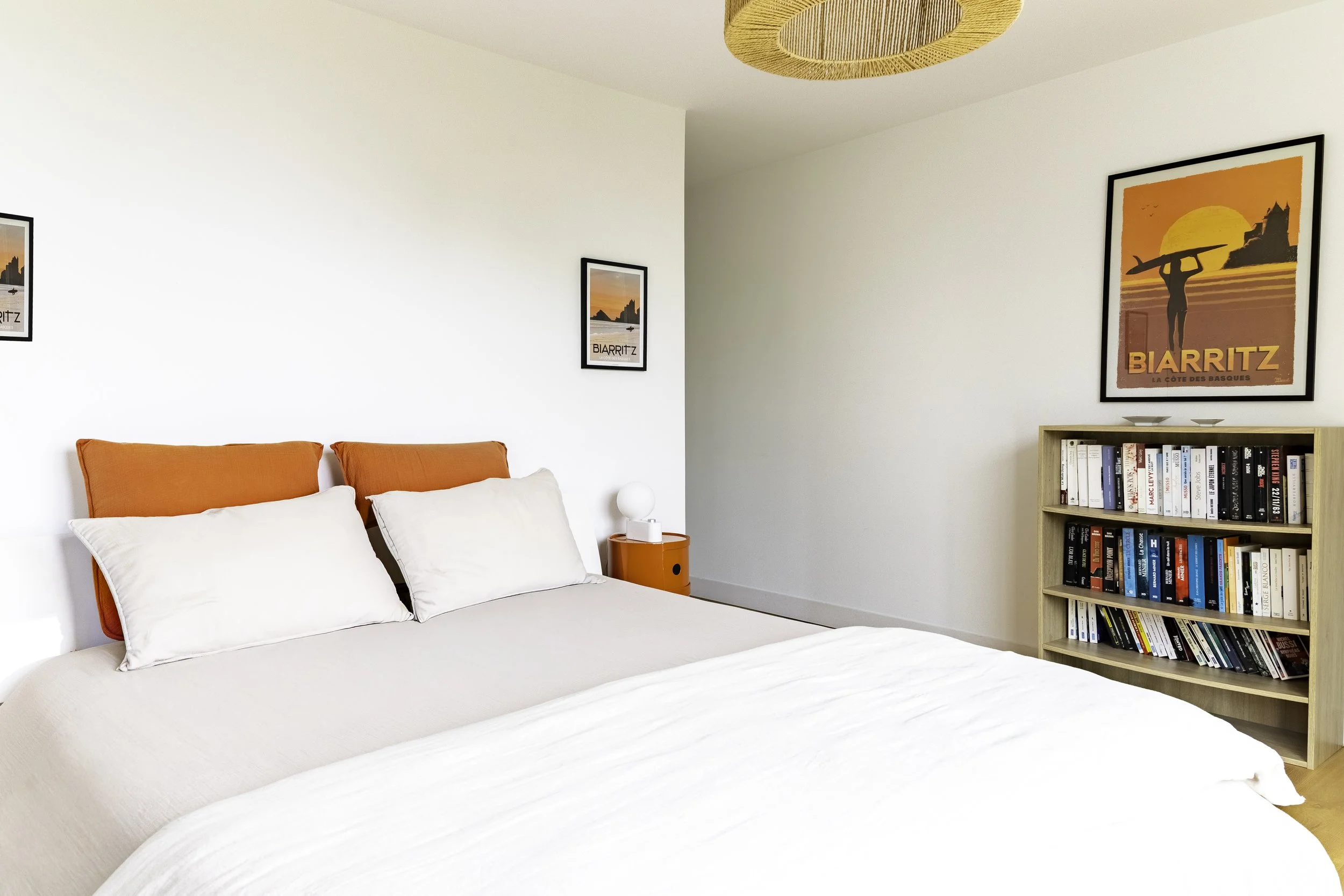 Chambre moderne avec un lit aux oreillers blancs et deux coussins orange, un fauteuil orange, une lampe de chevet blanche, et un mur décoré de posters dans un style vintage. Une étagère en bois contenant des livres et une décoration.