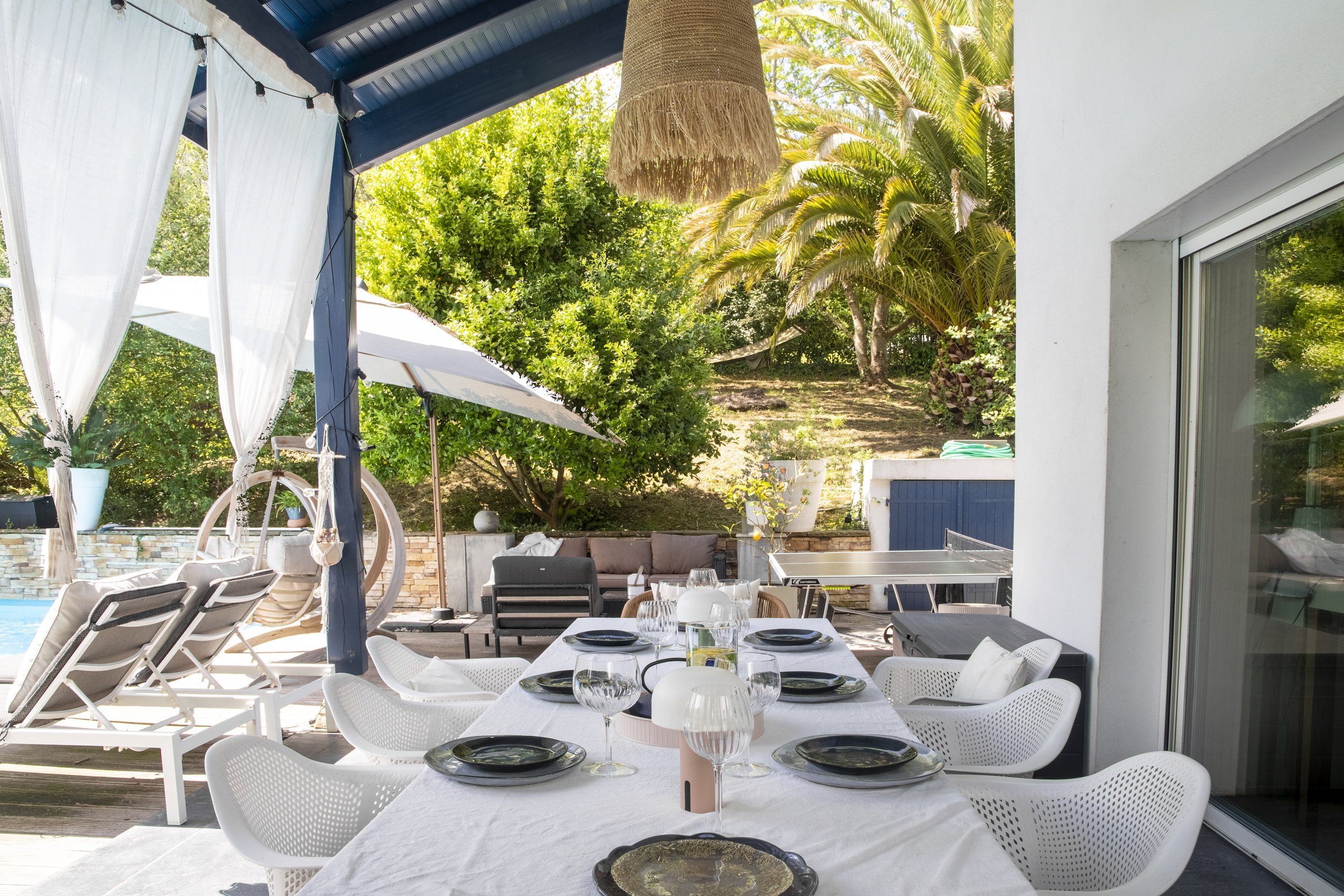 Une terrasse extérieure avec une table de manger prête pour un repas, entourée de chaises blanches, avec un décor végétal luxuriant et une maison moderne en arrière-plan.