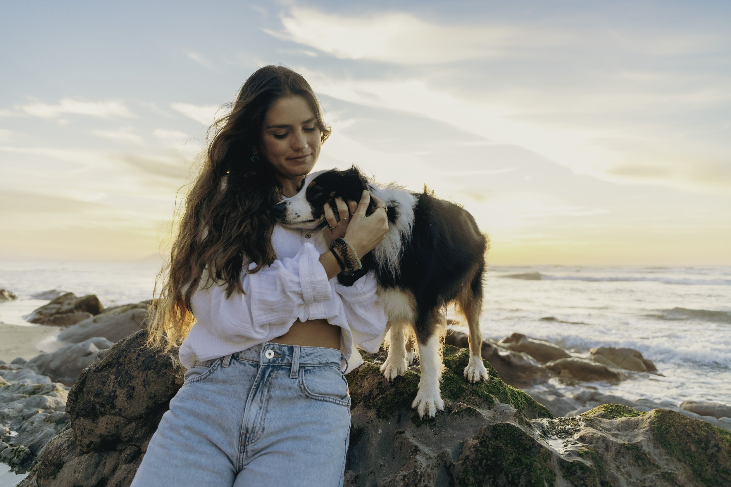Une jeune femme avec de longs cheveux bruns, portant un haut blanc et un jean, qui tient un chien Berner sur une roche au bord de la mer au coucher du soleil.