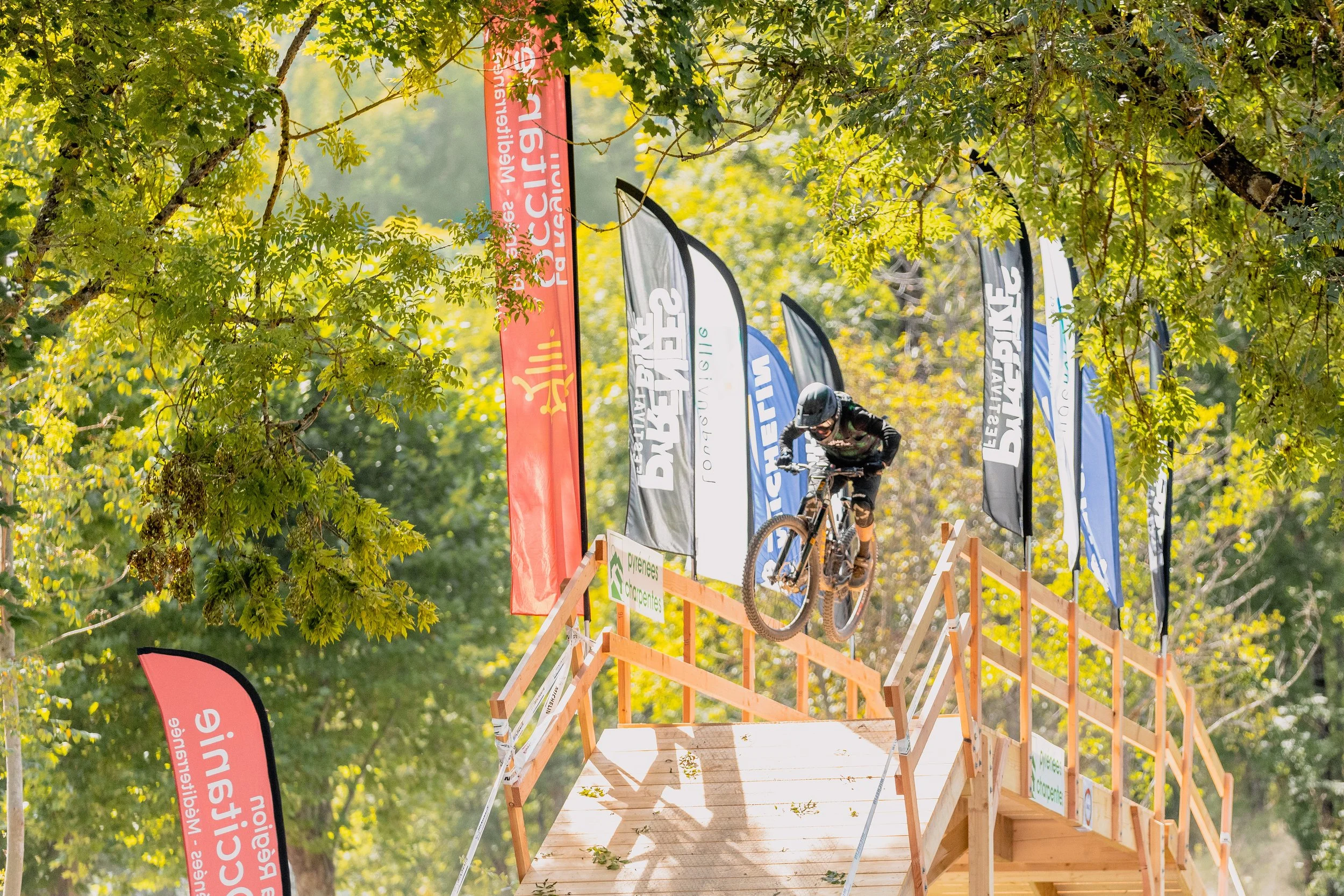 VTT (vélo tout terrain) en train de descendre une rampe en bois lors d'une compétition en plein air, entourée d'arbres avec des drapeaux publicitaires colorés.