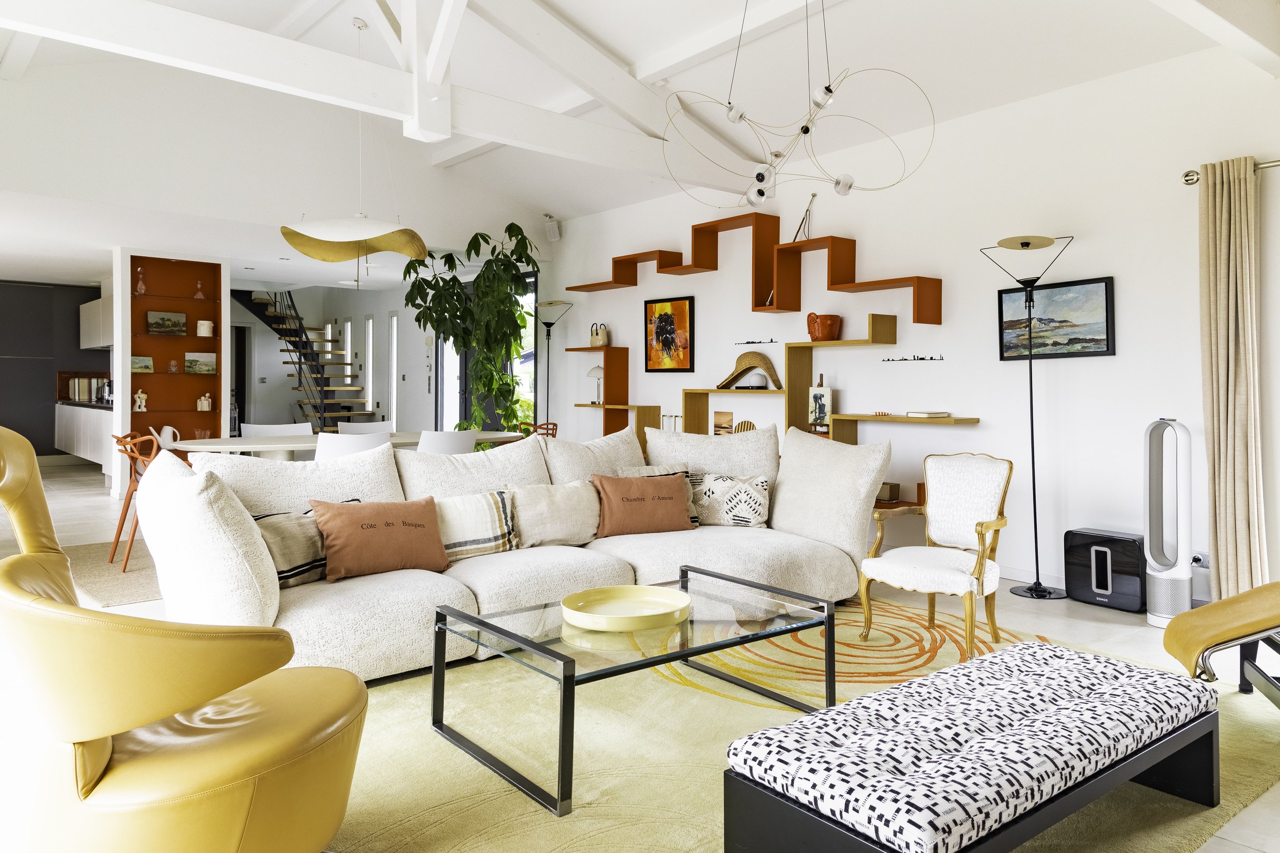 Salon moderne avec canapé beige, fauteuil jaune, table basse en verre, étagères rouges et jaunes, meubles, décoration artistique, plantes vertes, lampe suspendue, tableaux, tapis rayé orange et rose, enceinte audio et purificateur d'air