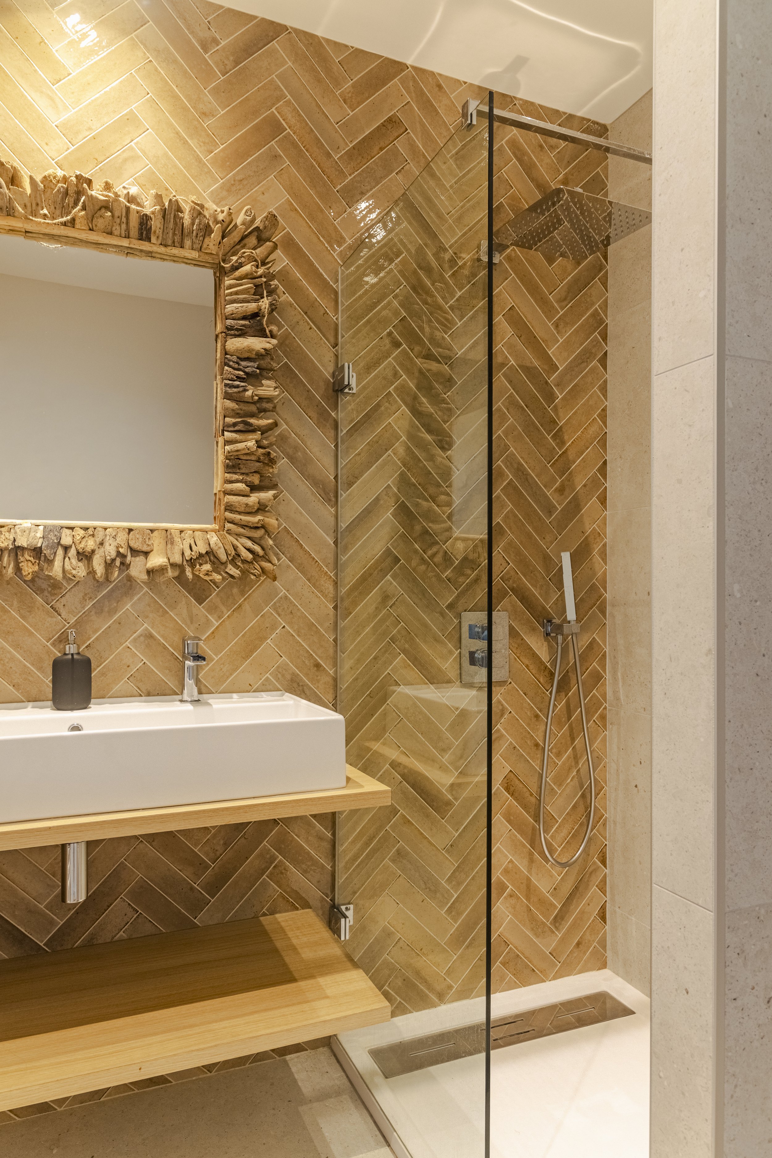 Salle de bain avec douche à l'italienne en verre, mur en tuiles de couleur beige disposées en motif en chevron, miroir encadré en bois, lavabo blanc sur une étagère en bois, un distributeur de savon noir sur le lavabo, et un sol en carrelage clair.