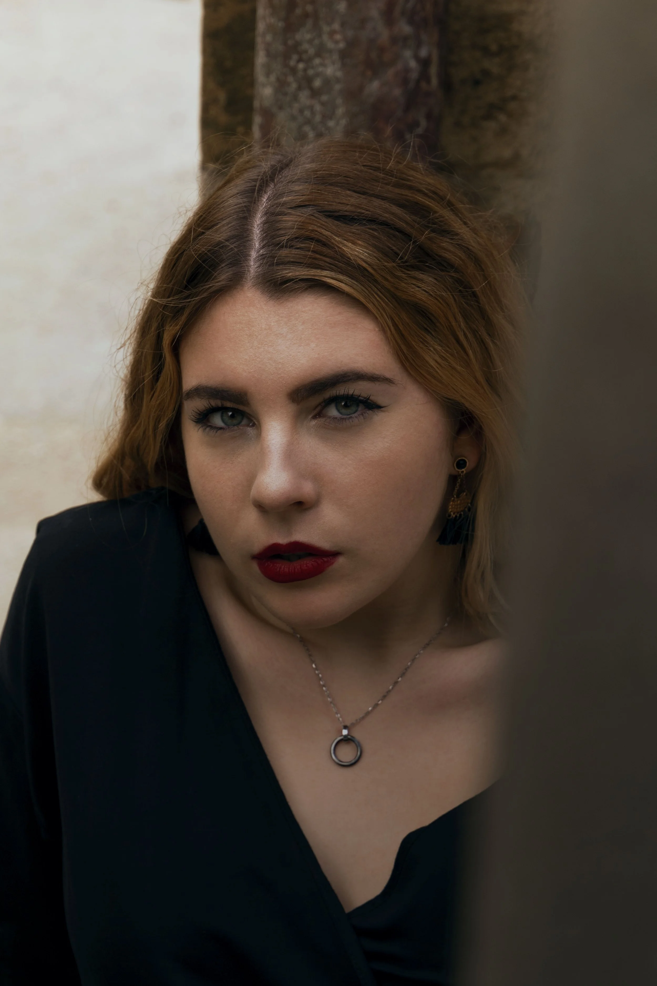 Jeune femme aux cheveux châtains, yeux clairs, maquillage sophistiqué, portant un collier et des boucles d'oreilles, posant contre un arbre en extérieur.