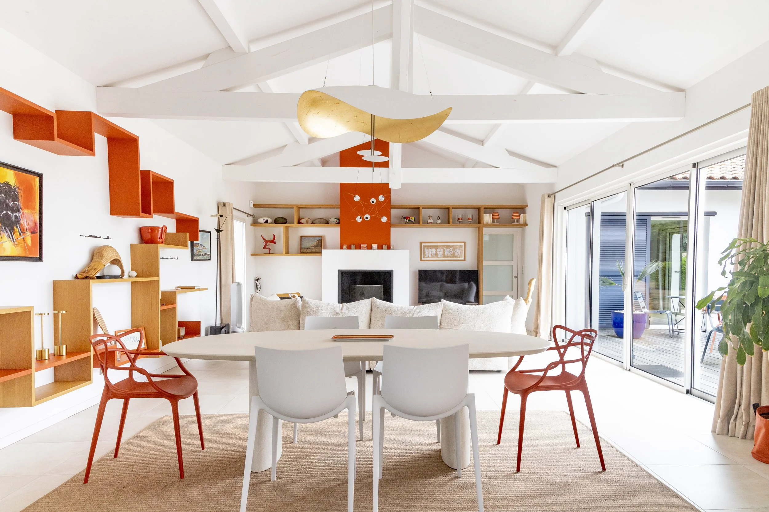Salle à manger avec table blanche, chaises rouges, étagères en bois, cheminée, grandes fenêtres avec rideaux, décoration colorée et moderne.