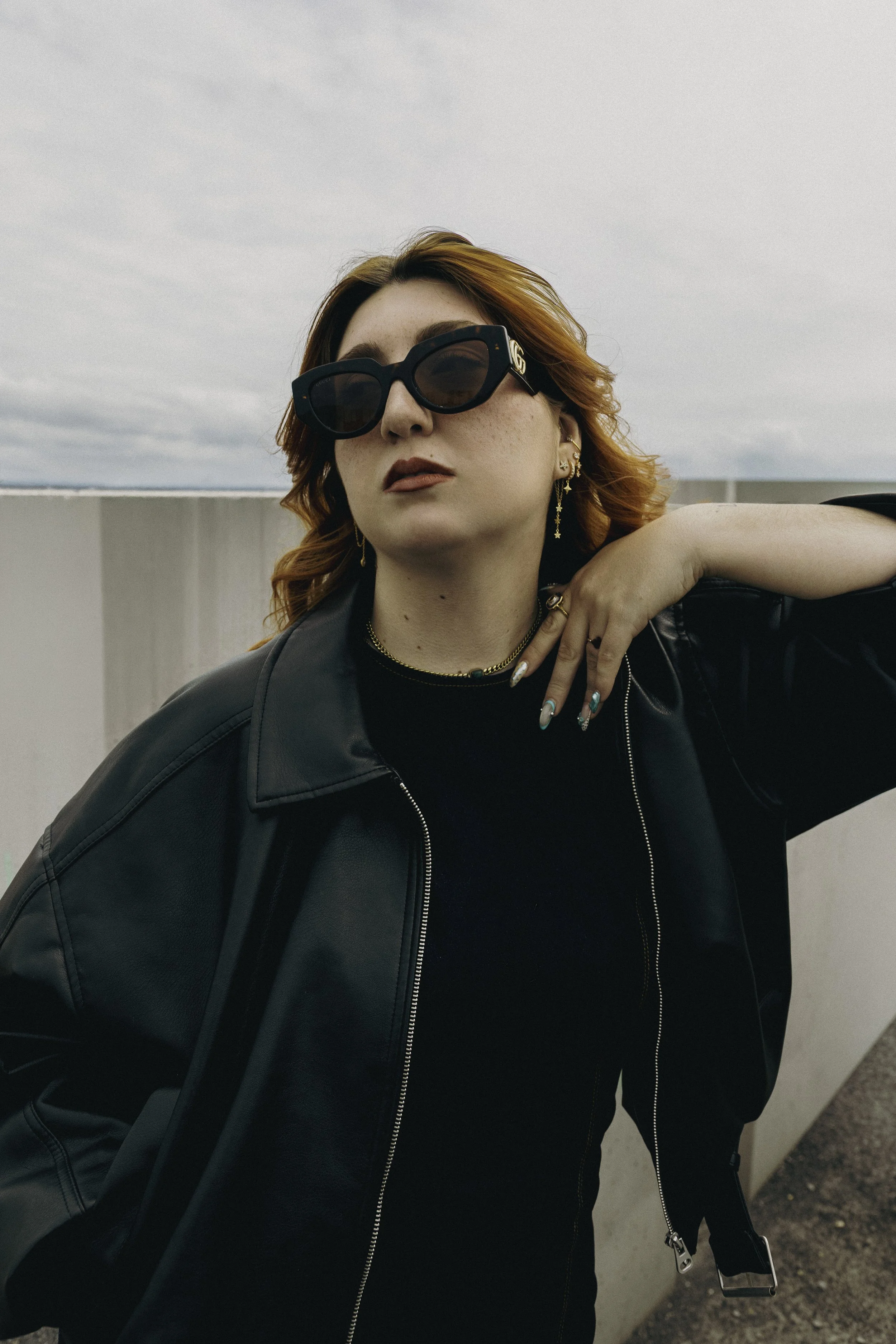Jeune femme avec cheveux roux, portant de grandes lunettes de soleil noires, des boucles d'oreilles pendantes dorées, une chaîne, un col roulé noir et une veste en cuir noir, posant avec la main sur son col, en extérieur contre un mur en béton et un ciel nuageux.