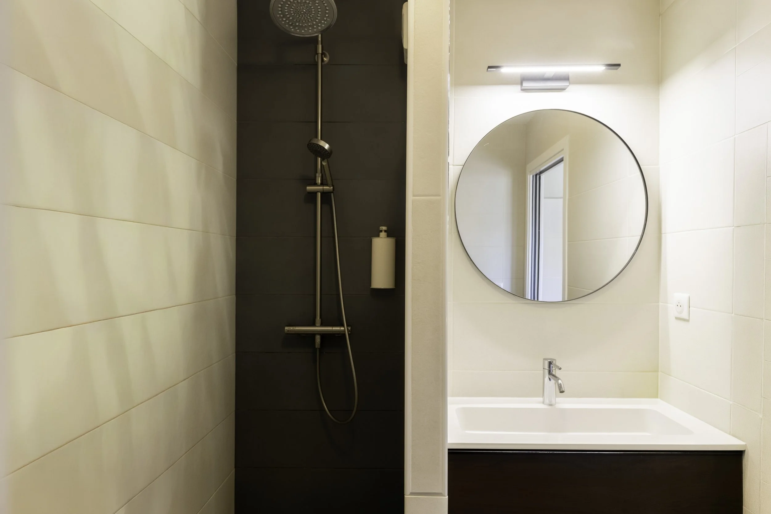 Une salle de bain avec une douche à gauche et une baignoire blanche à droite, un miroir rond au-dessus du lavabo.