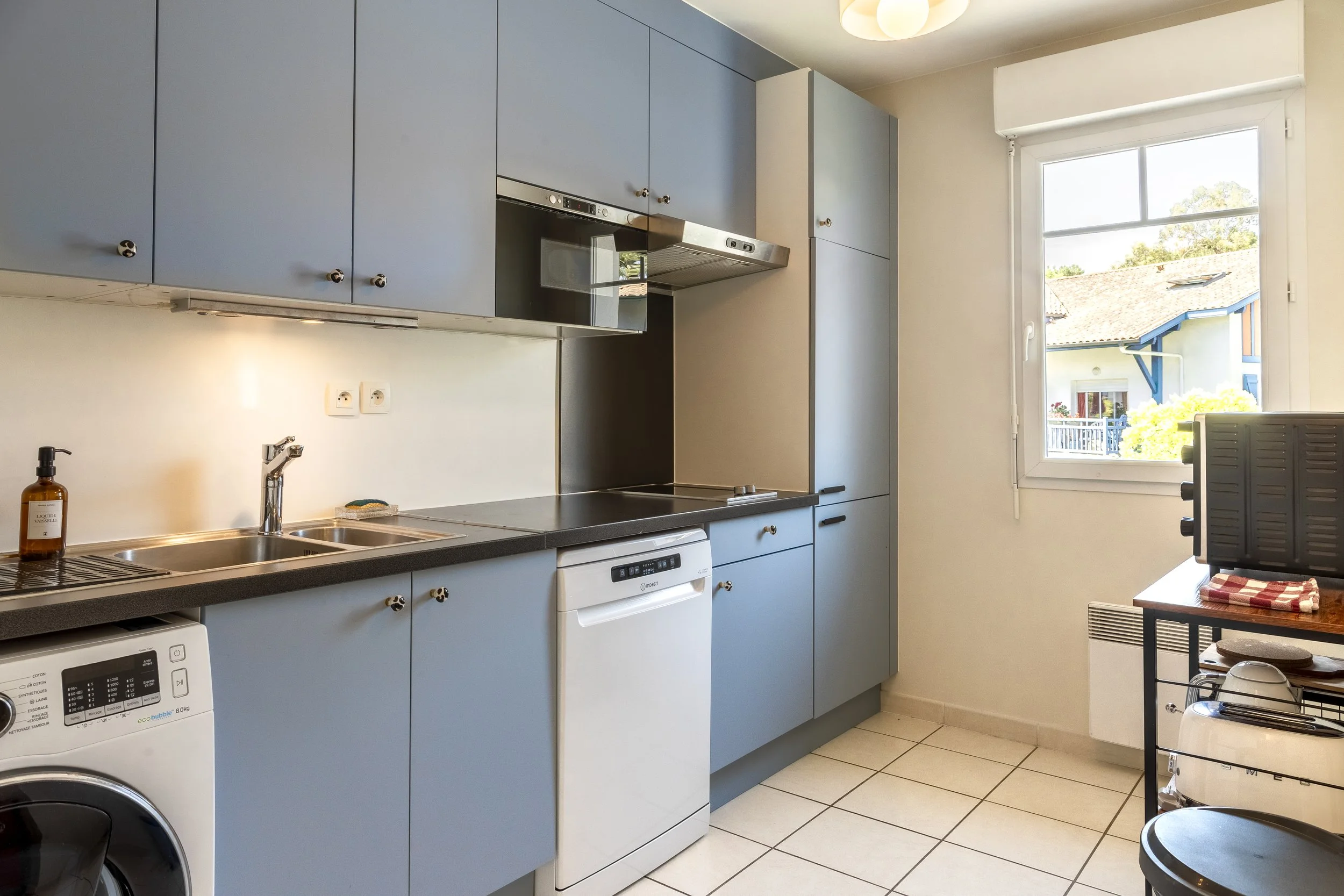 Cuisine avec des placards bleus, évier, lave-linge, four, réfrigérateur, une fenêtre avec vue sur une maison et des arbres, et un petit meuble à droite avec un grille-pain et un torchon à carreaux.
