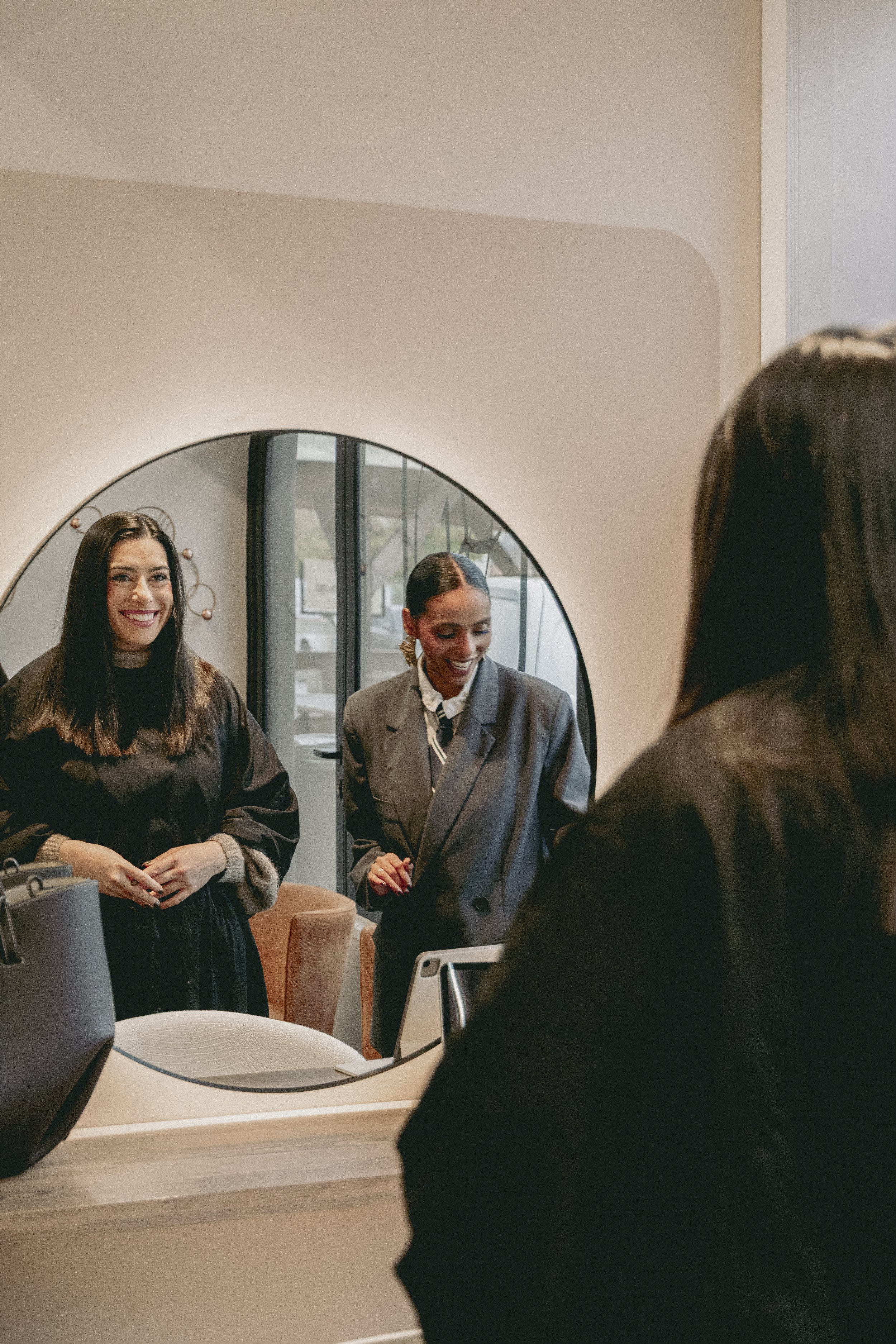 Trois femmes souriantes dans un espace intérieur, regardant un miroir, avec un décor moderne et des textiles confortables.