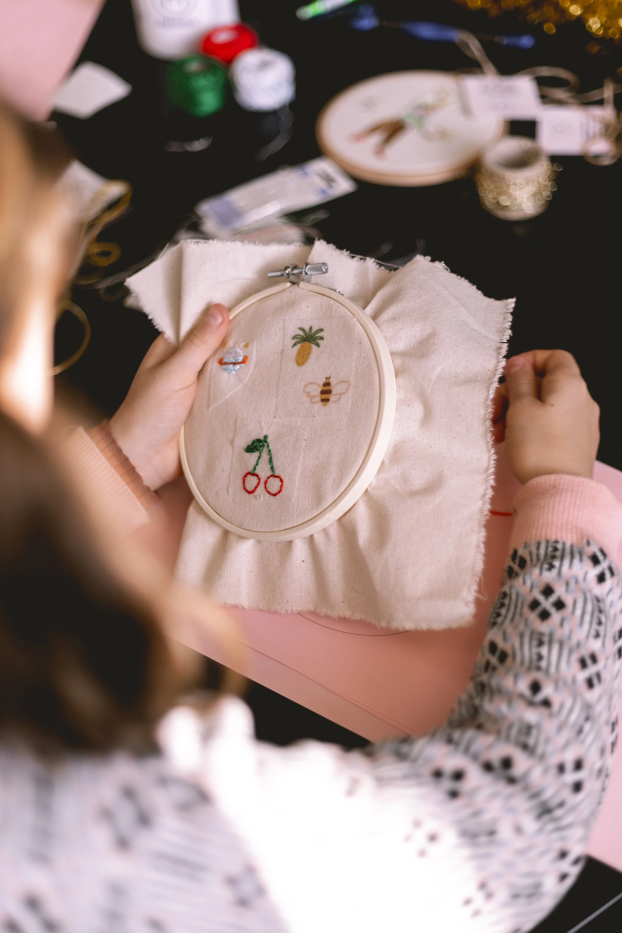 Fille tenant une pièce de tissu avec des broderies d'ananas, abeille, cerises et autres motifs, entourée d'outils de broderie et de décoration.