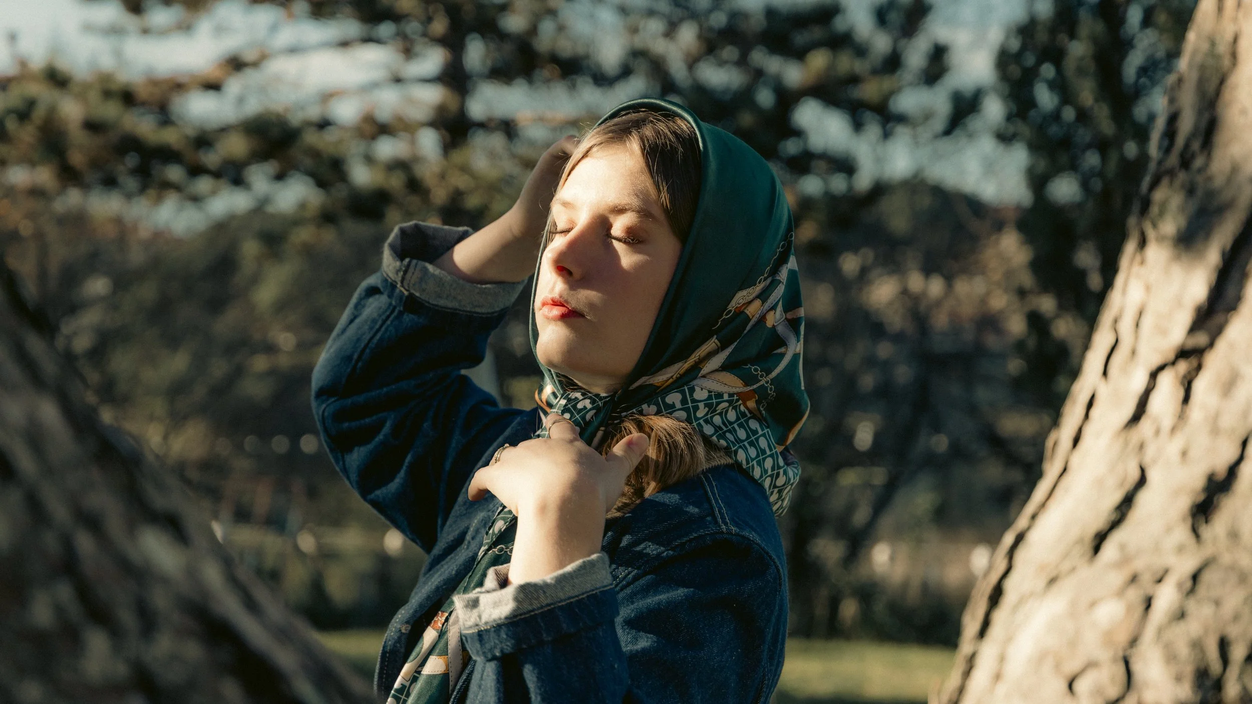 Jeune femme avec un foulard sur la tête, yeux fermés, profiter du soleil dans un parc en arrière-plan.