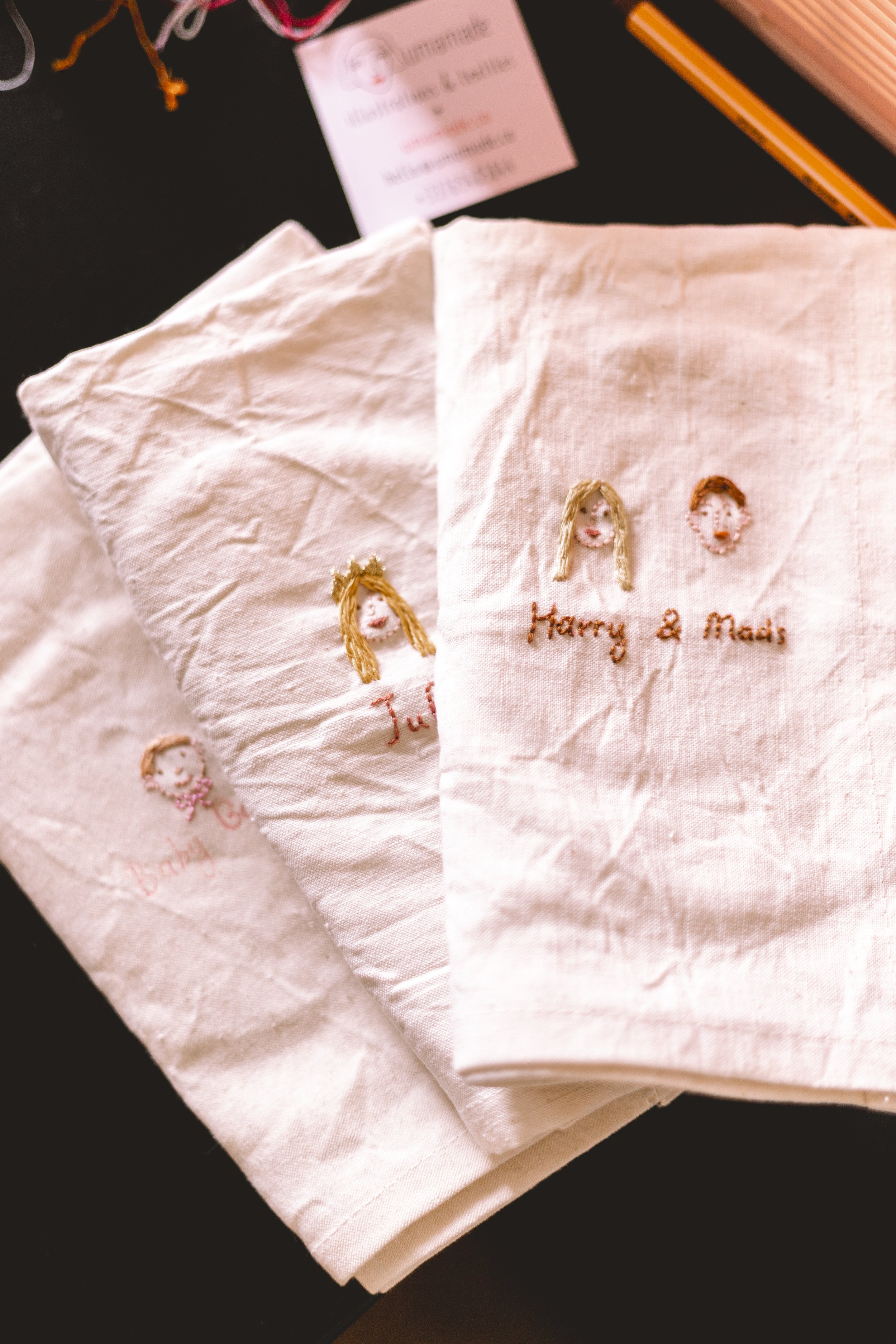 Des serviettes en tissu blanc avec une broderie représentant des visages de femmes et le texte 'Harry & Mads'.