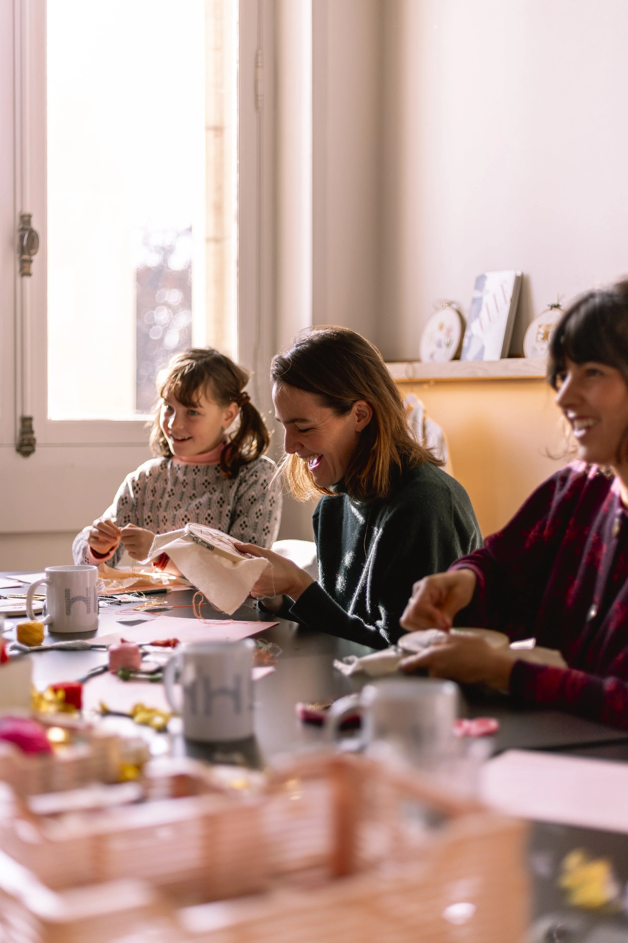 Groupe de trois personnes souriantes assises autour d'une table lors d'une activité créative, avec des fournitures de couture ou de broderie devant elles.
