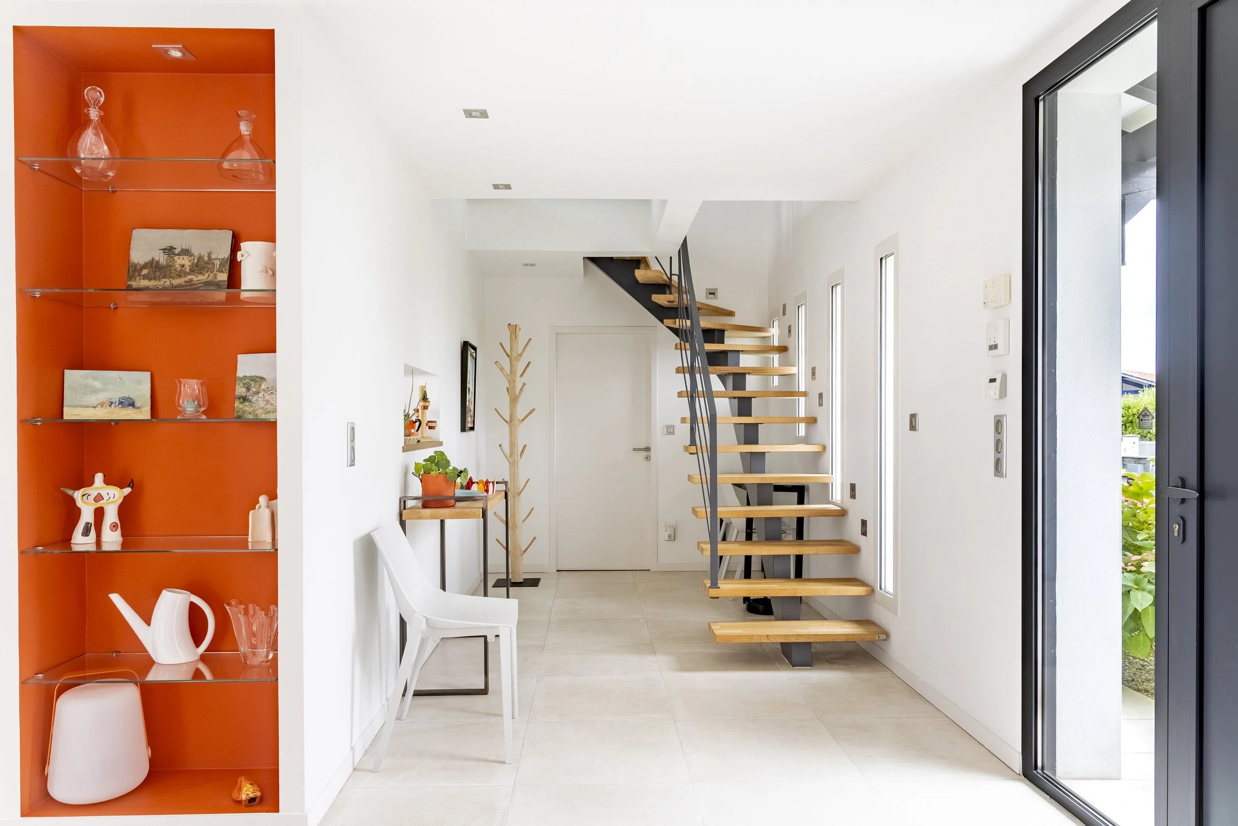 Intérieur d'une maison moderne avec escaliers en bois et murs blancs, étagère orange avec objets en verre et céramique, porte-fenêtre donnant sur le jardin.