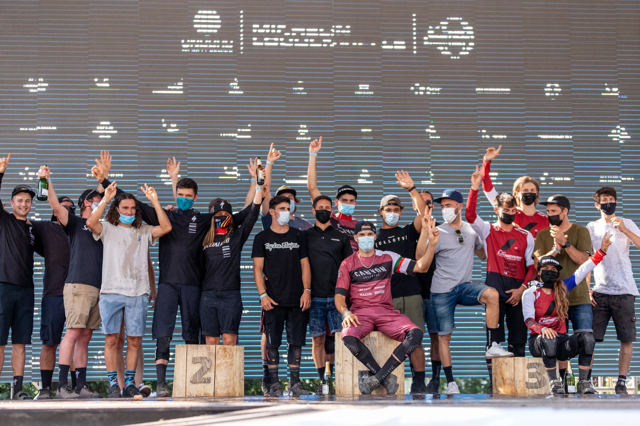 Groupe de cyclistes sur un podium, certains tenant des bouteilles de boissons, portant des maillots de course, certains portant des masques faciaux, en célébration de leur victoire, avec un grand écran numérique en arrière-plan.