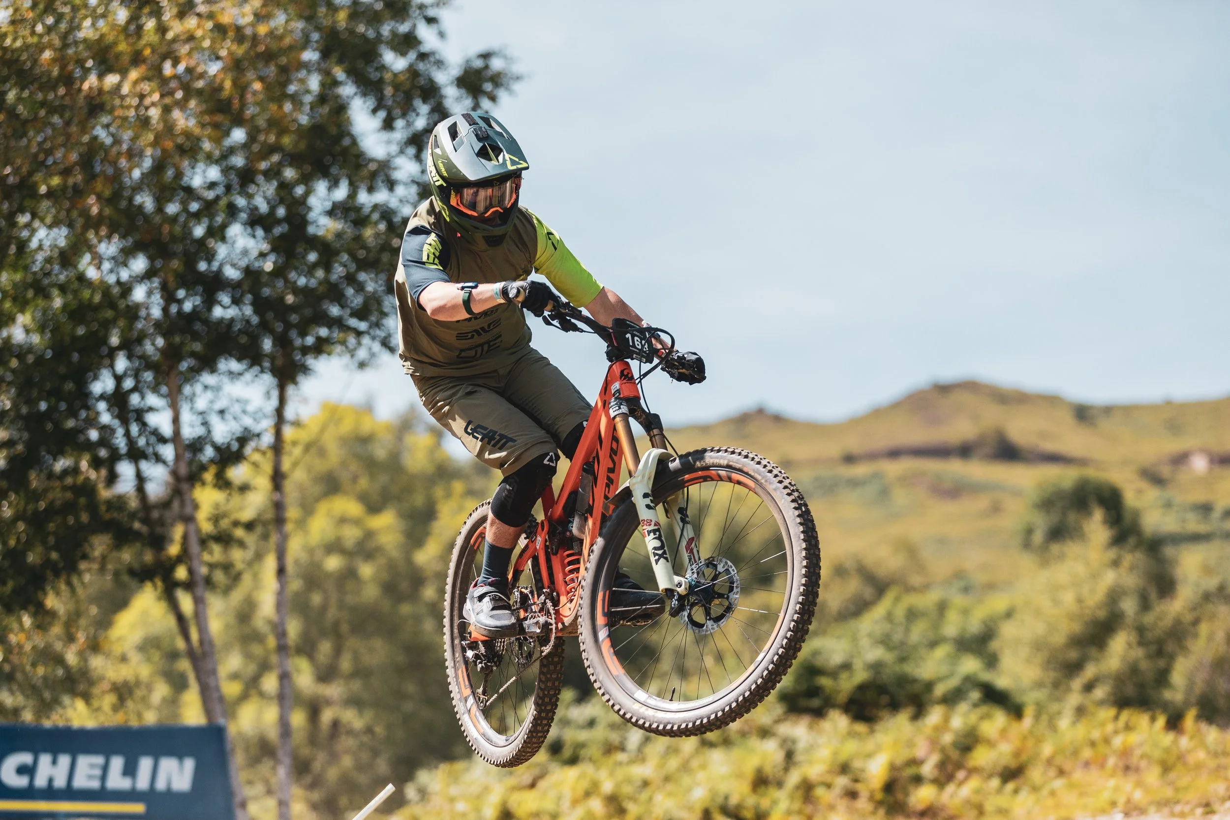 Vélo tout-terrain en plein saut avec un cycliste portant un casque et une tenue de sport, dans un paysage naturel avec des arbres et des collines en arrière-plan.