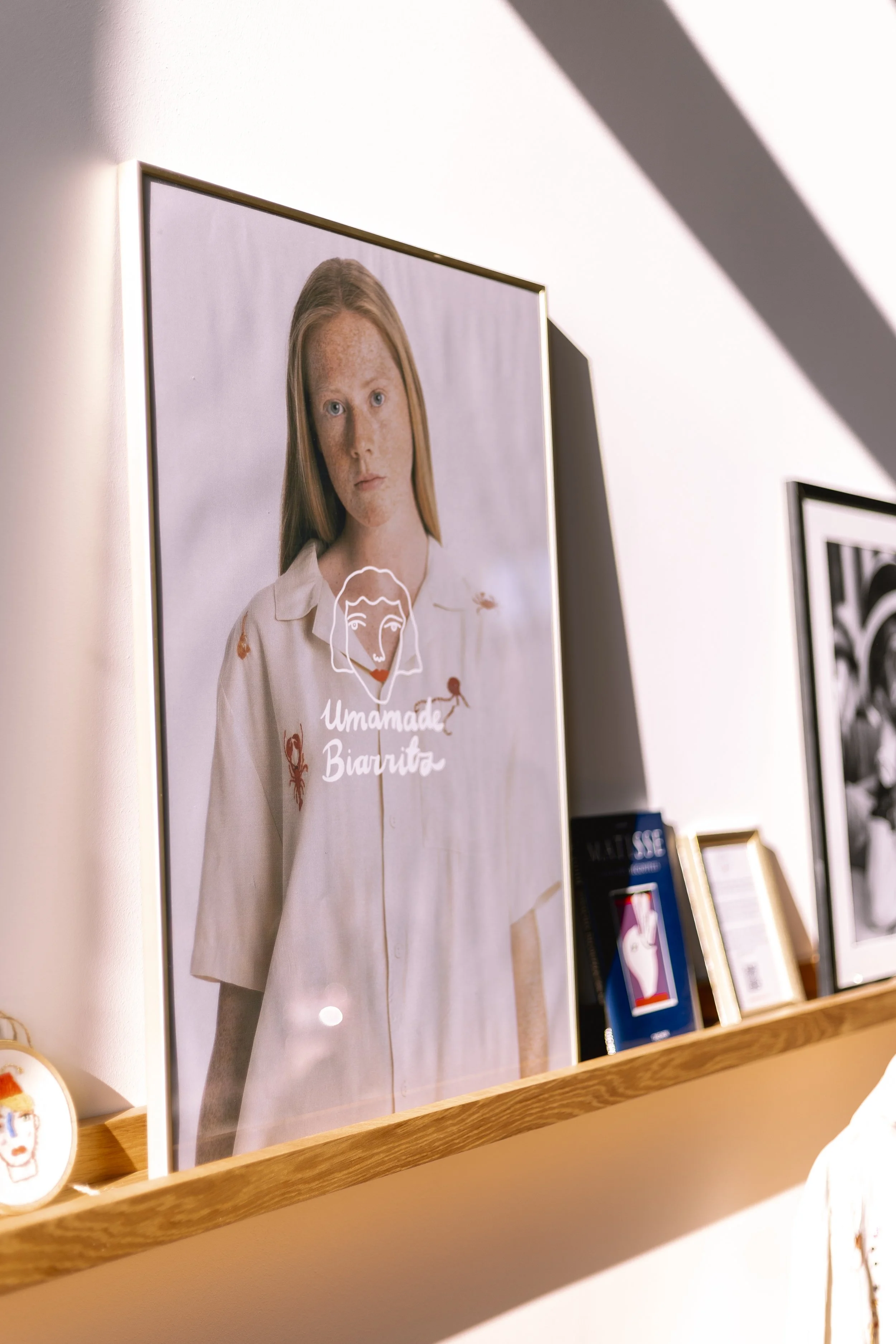 Affiche avec un portrait d'une jeune femme rousse aux taches de rousseur, portant une chemise blanche, avec le texte "Unmade Biarritz" dessous, affichée sur un mur blanc dans une galerie d'art, avec des livres et autres œuvres en arrière-plan.