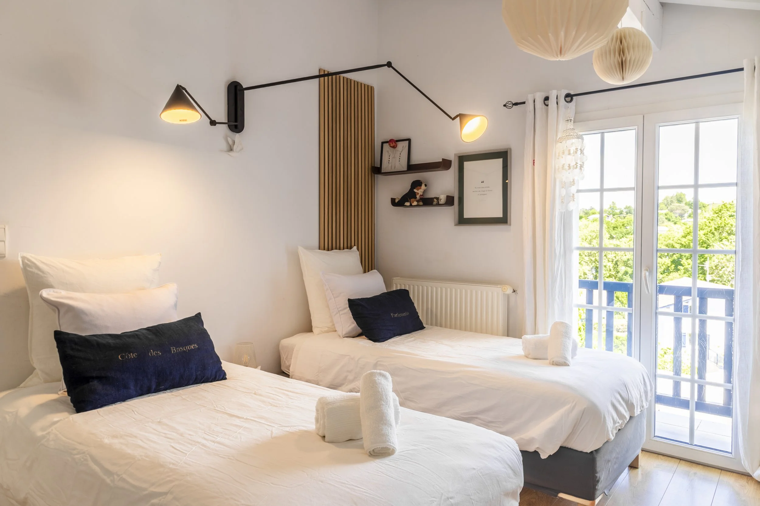 Chambre avec deux lits, draps blancs, oreillers avec housses bleues, décoration moderne, grande fenêtre avec rideaux blancs laissant entrer la lumière et montrant un paysage verdoyant extérieur, lumière murale réglable, lampes en papier suspendues au plafond.