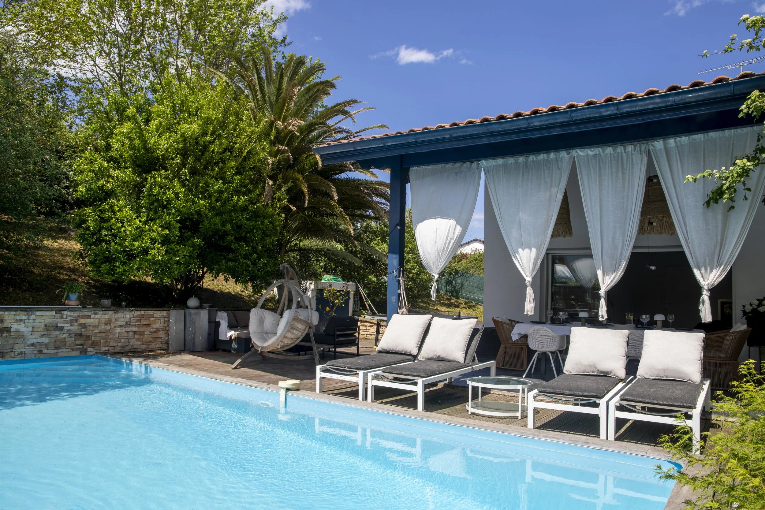 Une piscine extérieure avec des chaises longues blanches, un espace détente avec des coussins, une pergola avec des rideaux blancs, entourée de végétation luxuriante et un ciel bleu avec quelques nuages.