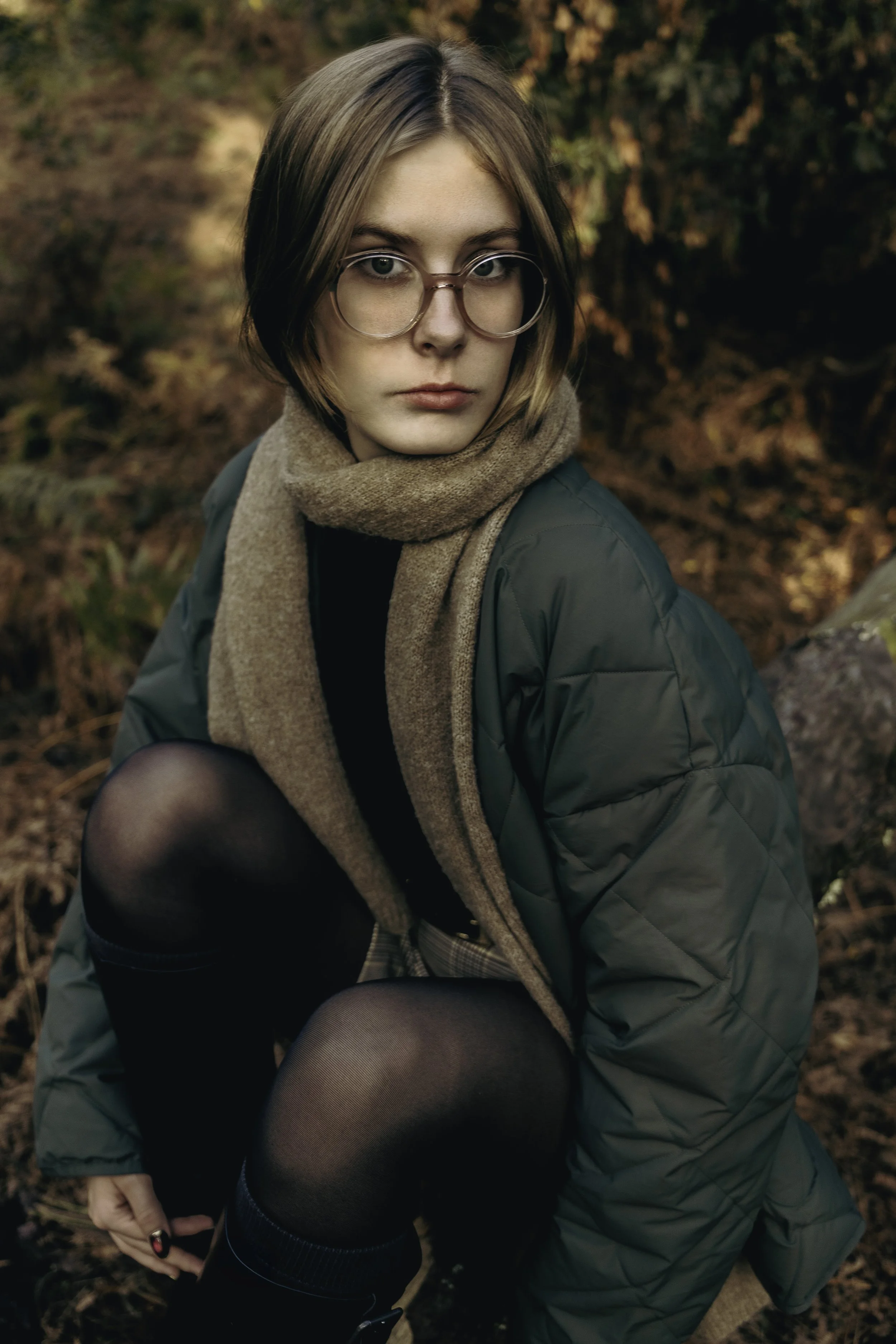 Une jeune femme avec des lunettes, vêtue d'un manteau vert et d'une écharpe beige, dans un environnement d'automne avec des feuilles orange et marron.