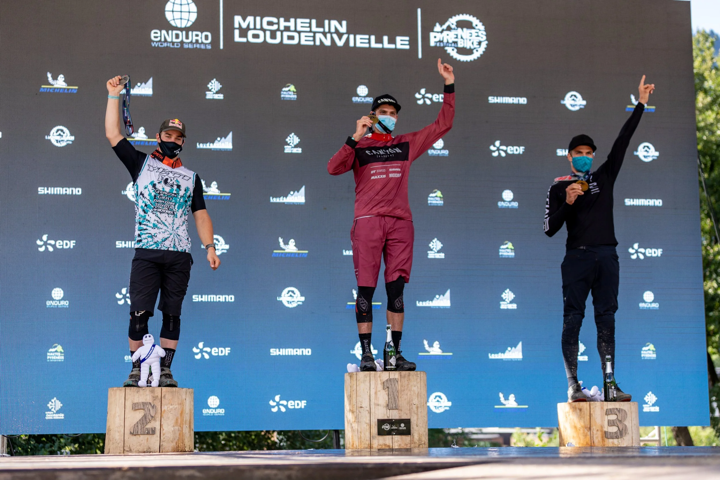 Trois cyclistes en tenue de compétition sur un podium en bois, recevant des médailles. L'arrière-plan comporte un panneau avec les logos Enduro World Series, Michelin Loudenvielle, et d'autres sponsors, avec des arbres visibles sur le côté droit.