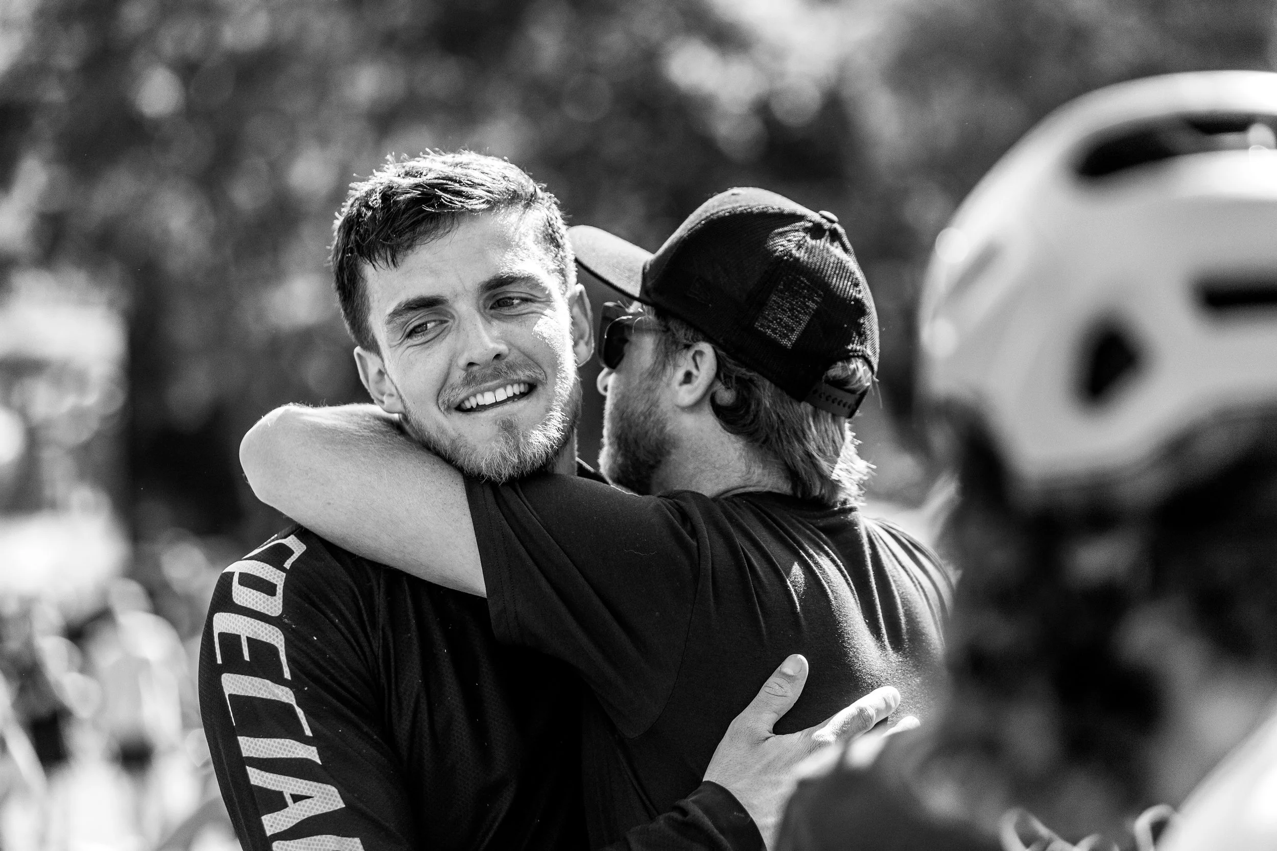 Deux hommes souriants se serrent dans une accolade lors d'une rencontre amicale en extérieur, en noir et blanc.