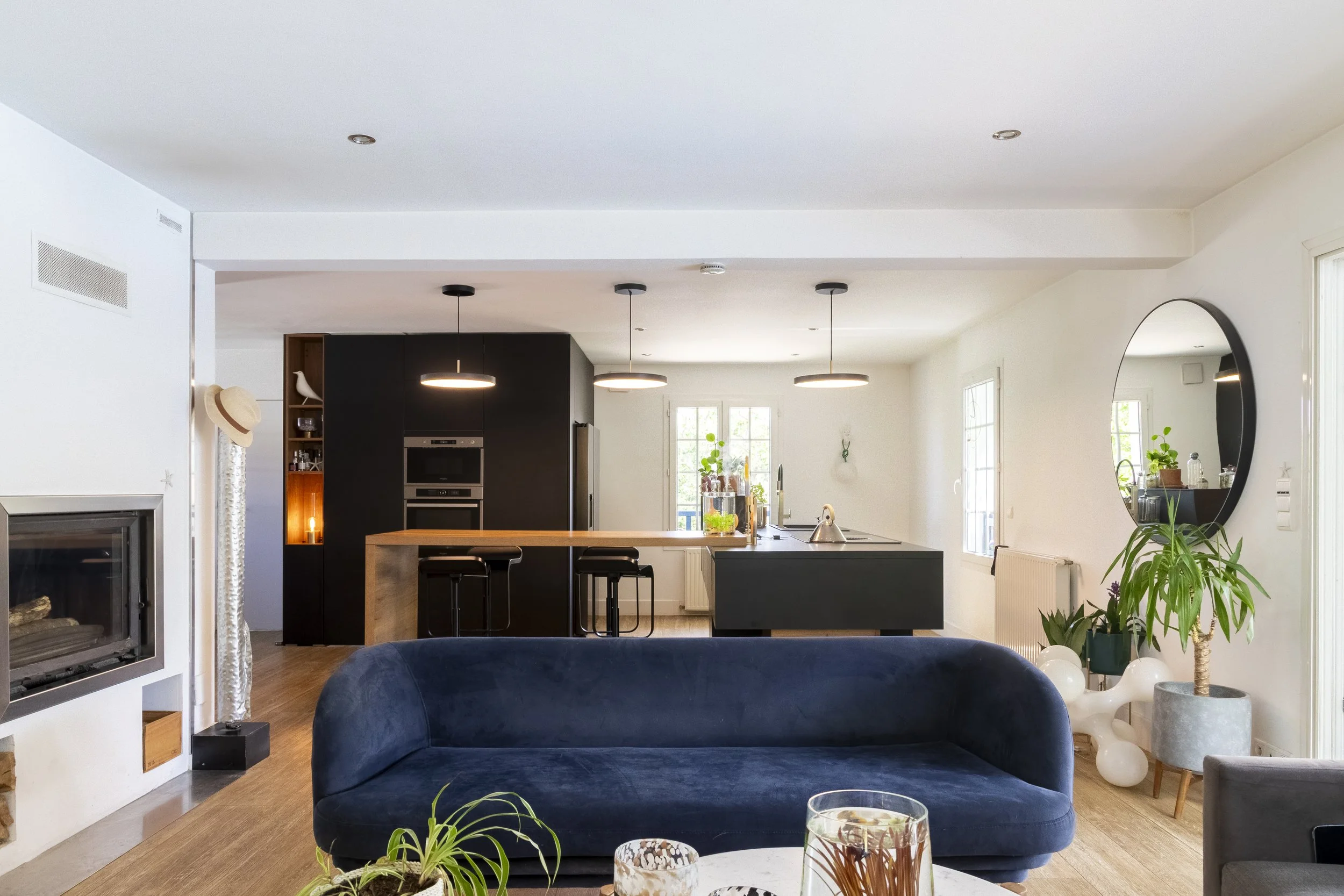 Intérieur moderne d'une cuisine ouverte, avec un îlot central en bois, des lampes suspendues, un miroir rond, des plantes vertes, et un canapé en velours bleu.