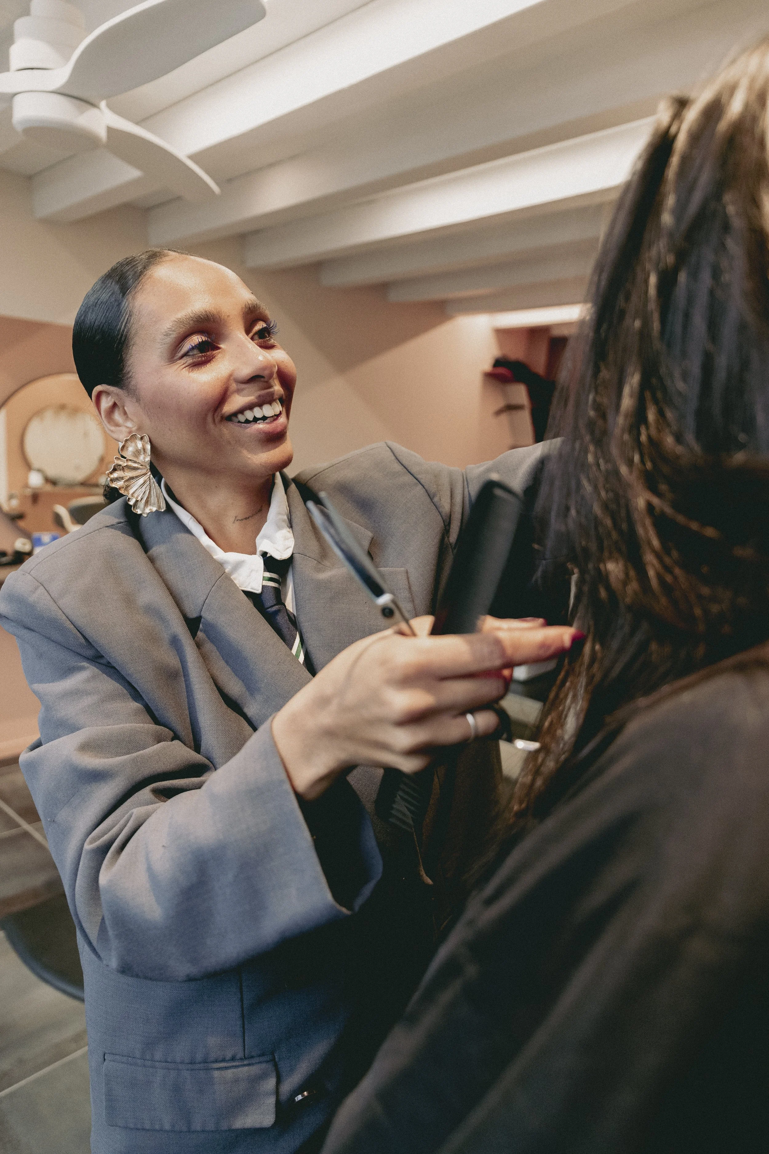 Une femme souriante en costume gris, portant une boucle d'oreille large en forme de coquillage en or, tient un micro lors d'une interview ou d'une conversation dans un intérieur