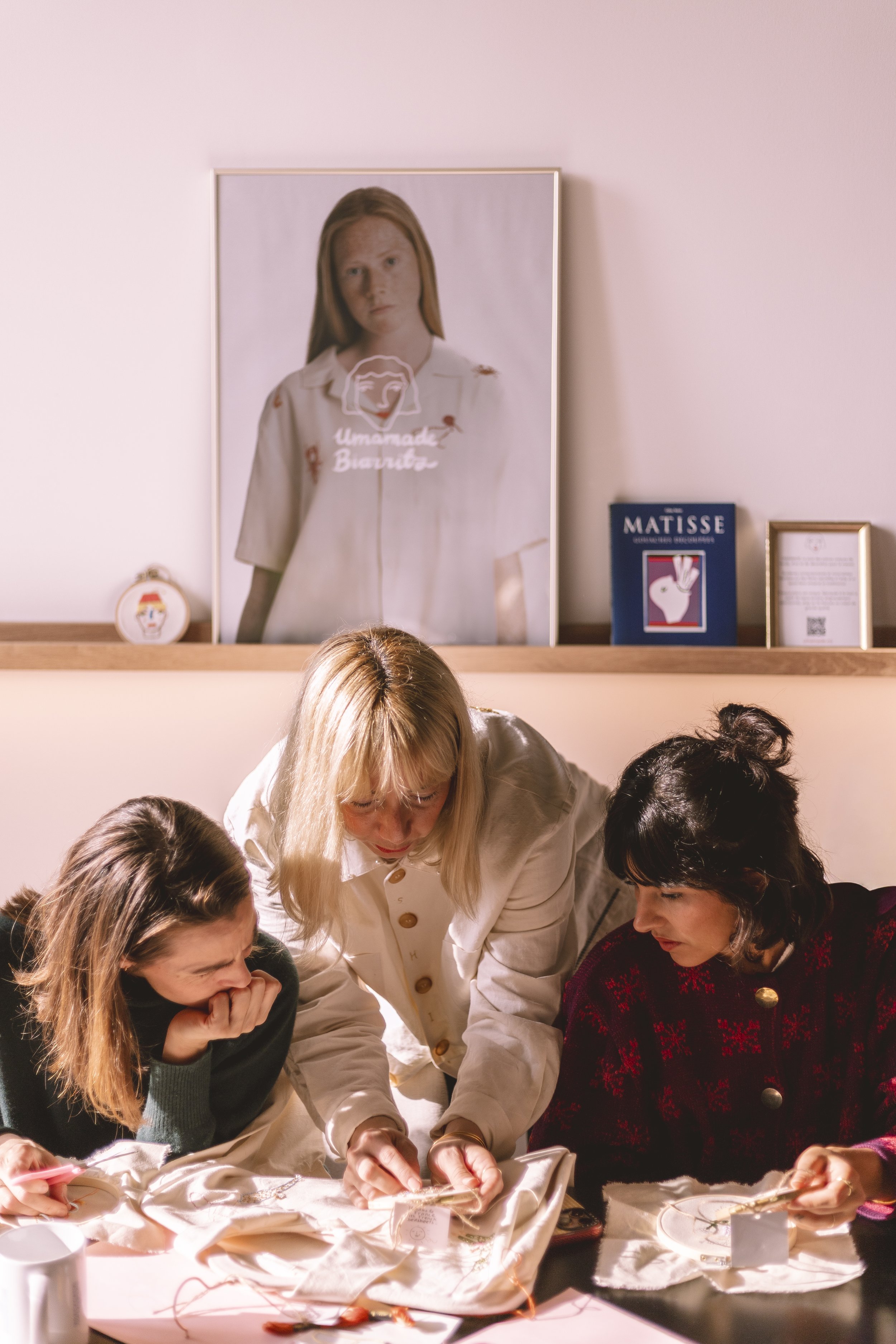 Un groupe de personnes, dont une femme blonde en manteau beige, travaille ensemble sur un projet sur une table, avec des décorations de Noël. En arrière-plan, poster d'une femme à cheveux rouges et livres d'art.