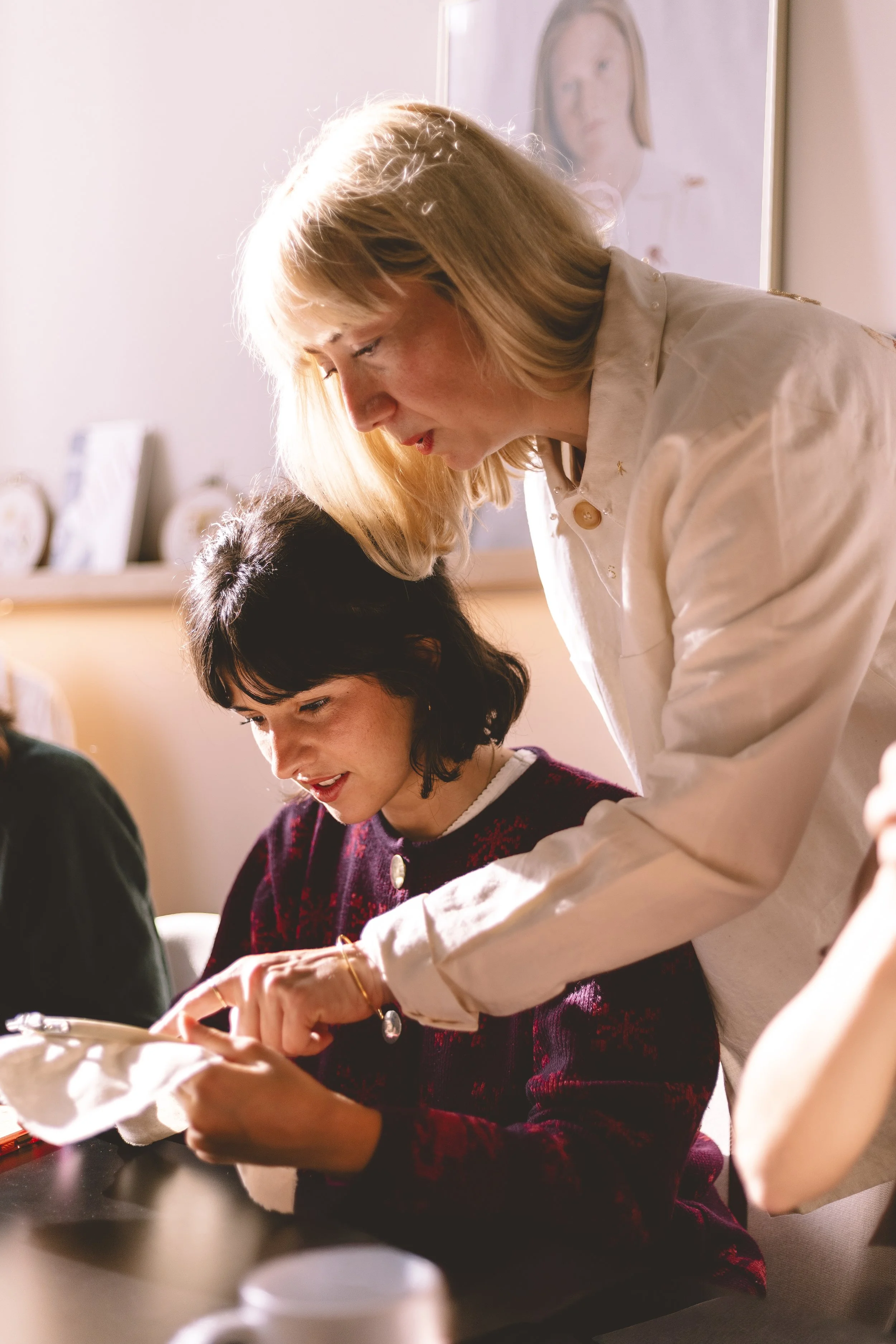 Une femme blonde et une femme avec des cheveux noirs travaillent ensemble autour d'une table, concentrées sur une activité, dans un intérieur chaleureux avec des décorations et des peintures en arrière-plan.