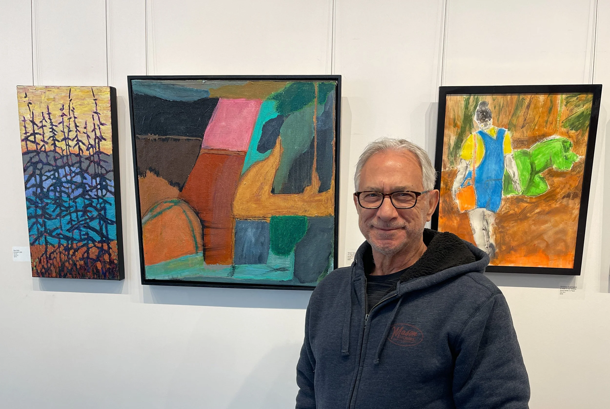 A man with gray hair, glasses, and a gray hoodie standing in front of three abstract paintings in an art gallery.