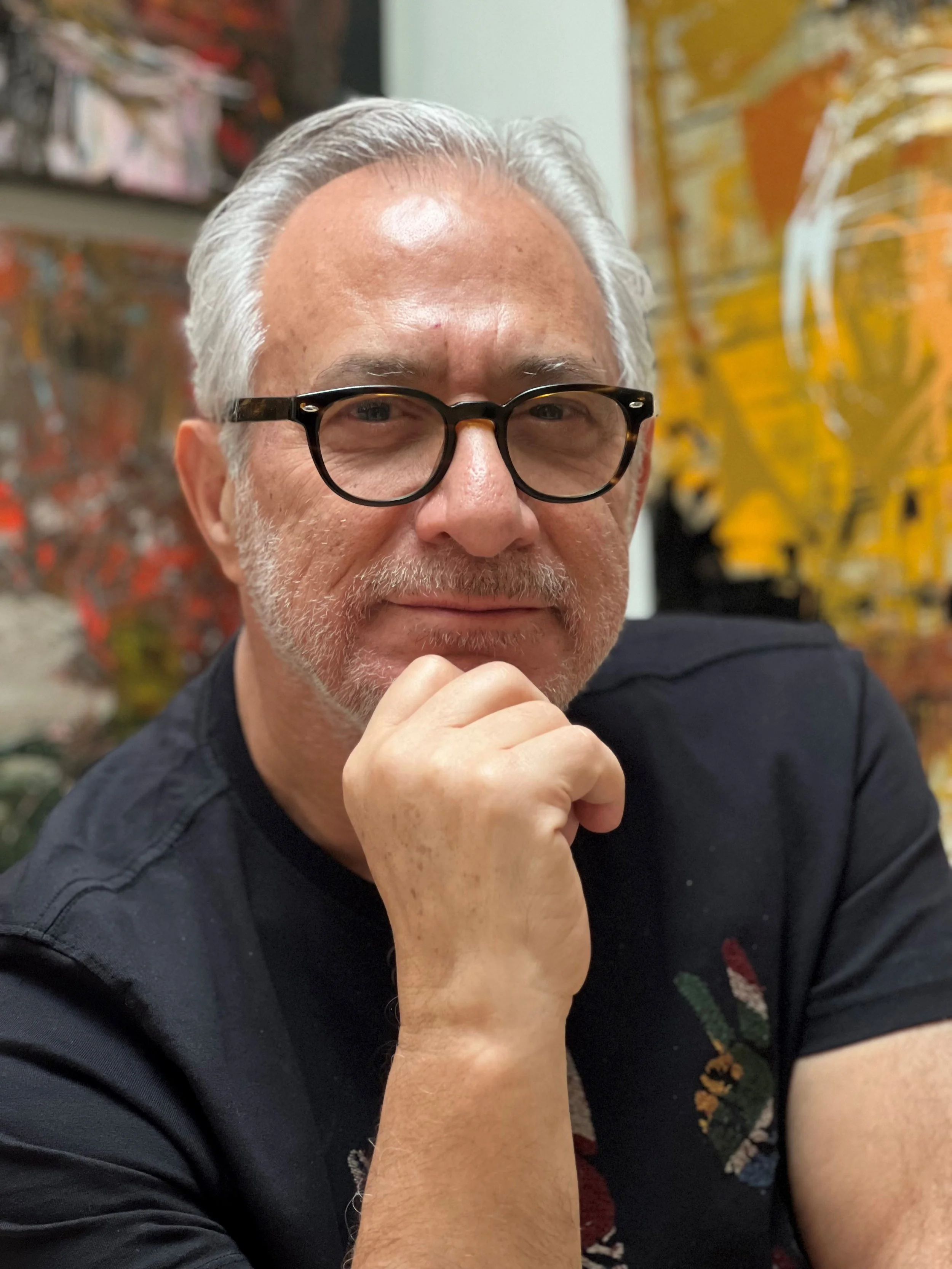 A middle-aged man with gray hair, glasses, and a beard, resting his chin on his hand and looking at the camera. He is wearing a black shirt with colorful embroidery on the sleeve. The background features colorful abstract artwork.