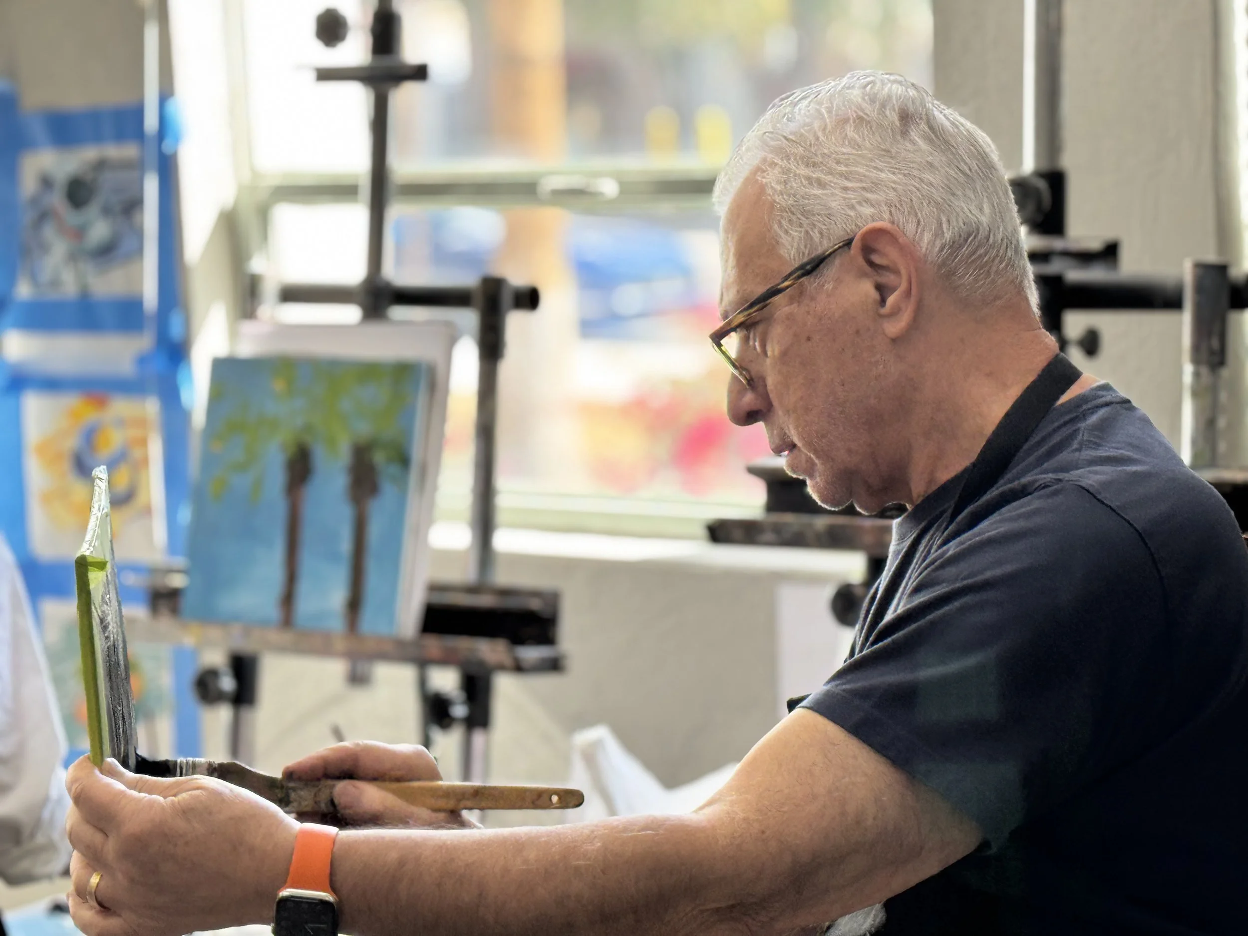 An elderly man with gray hair and glasses painting a picture of palm trees on a canvas in an art studio.