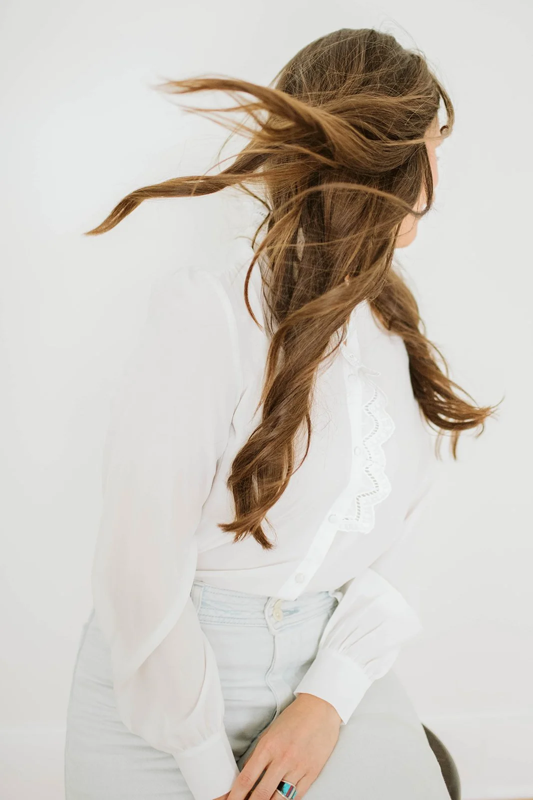 A woman with long, wavy brown hair, wearing a white shirt and light pants, sitting against a plain white background.