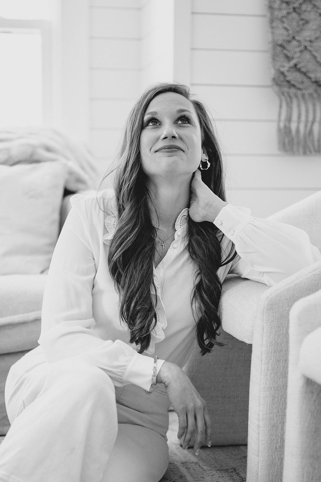 Black and white photo of a woman with long hair sitting on the floor, leaning on a sofa, looking upward with a gentle smile, in a cozy living room.