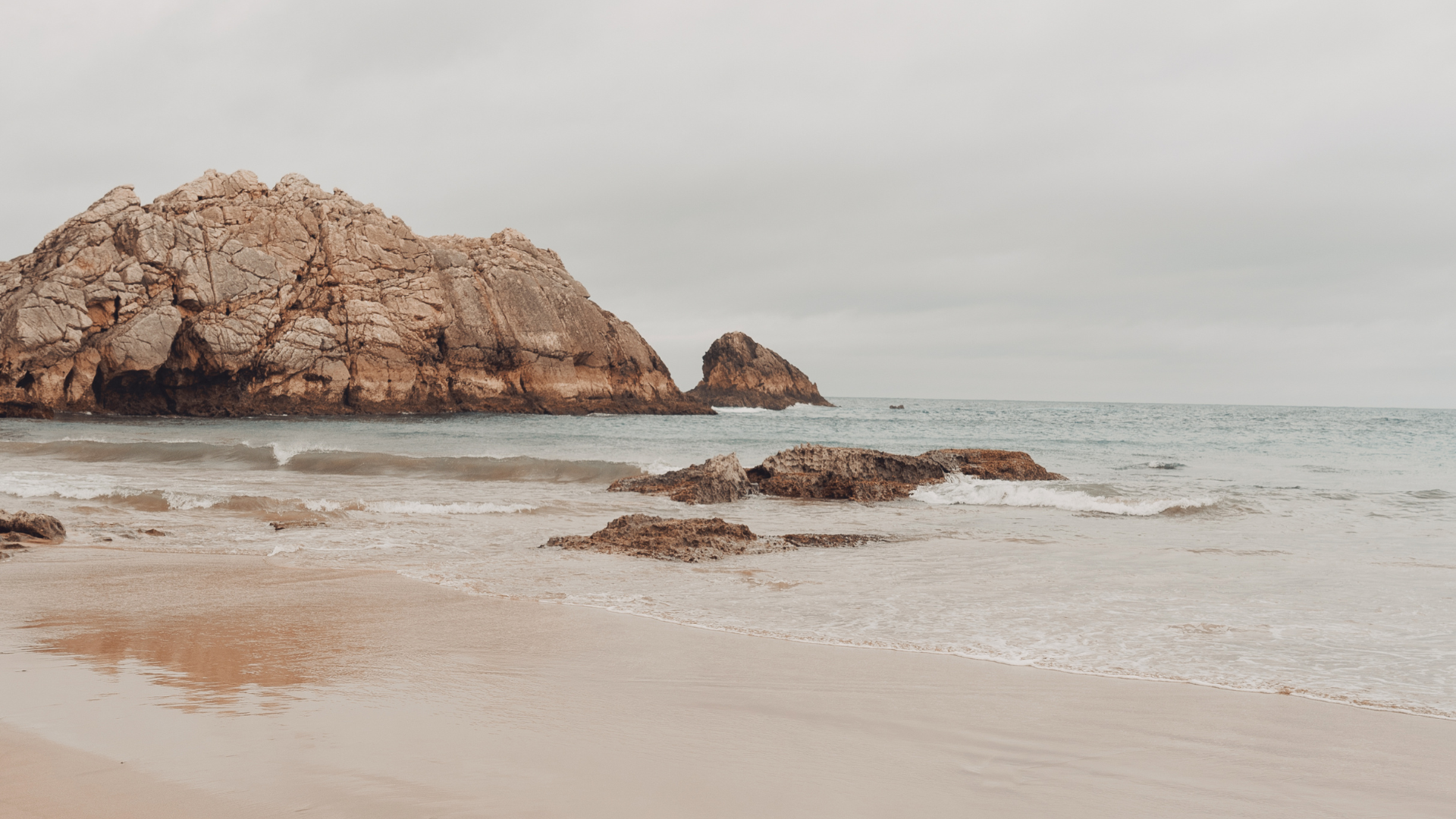 Scenic view of a beach with rocks in the foreground, gentle waves, a large rocky island or cliff in the background, and a cloudy sky.
