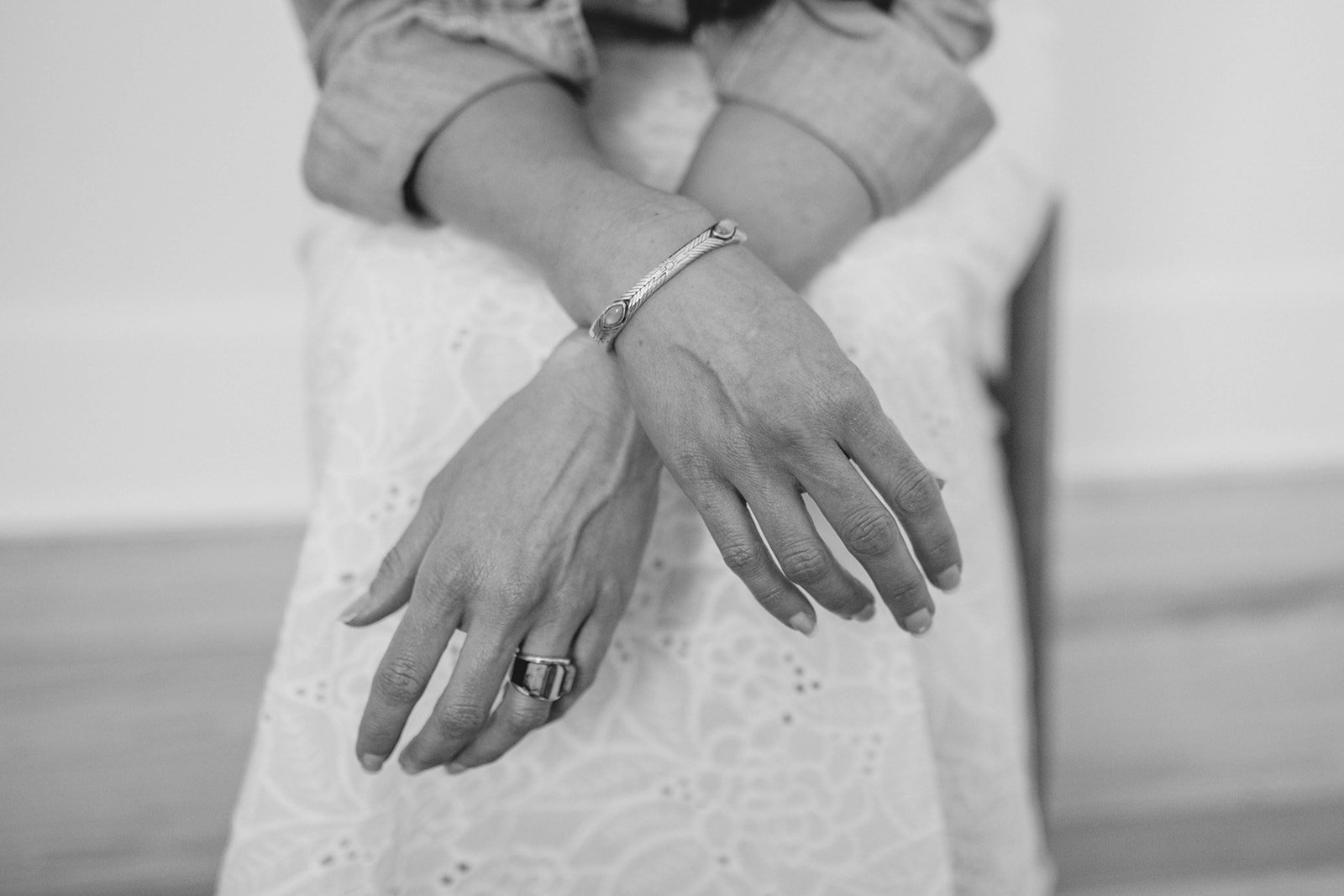 Close-up of a woman's hands crossed, wearing a bracelet on her wrist and a ring on her finger, with a lace dress in the background.