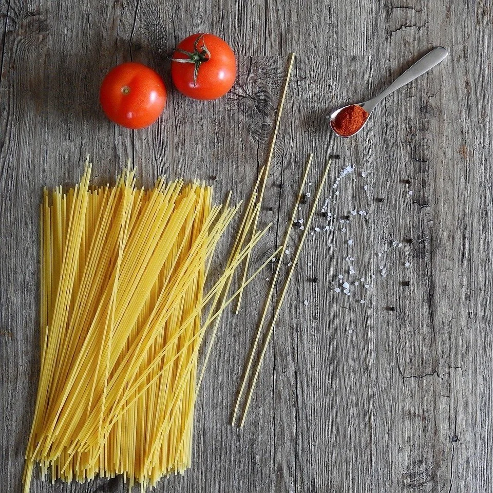 Des spaghettis crus, deux tomates, une cuillère de paprika en poudre, grains de poivre et sel sur une table en bois.