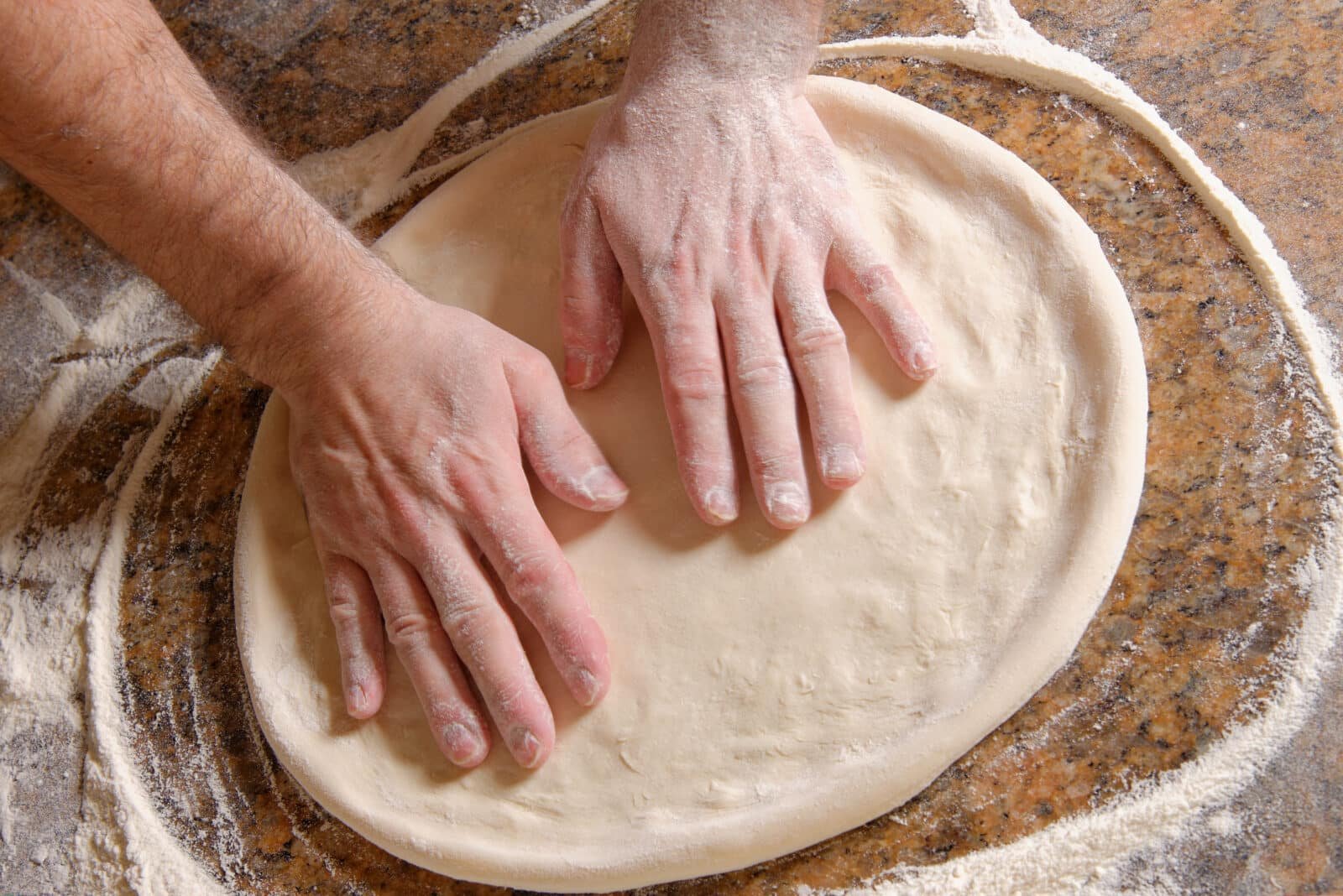 Mains étalant une pâte à pizza sur une surface farinée.